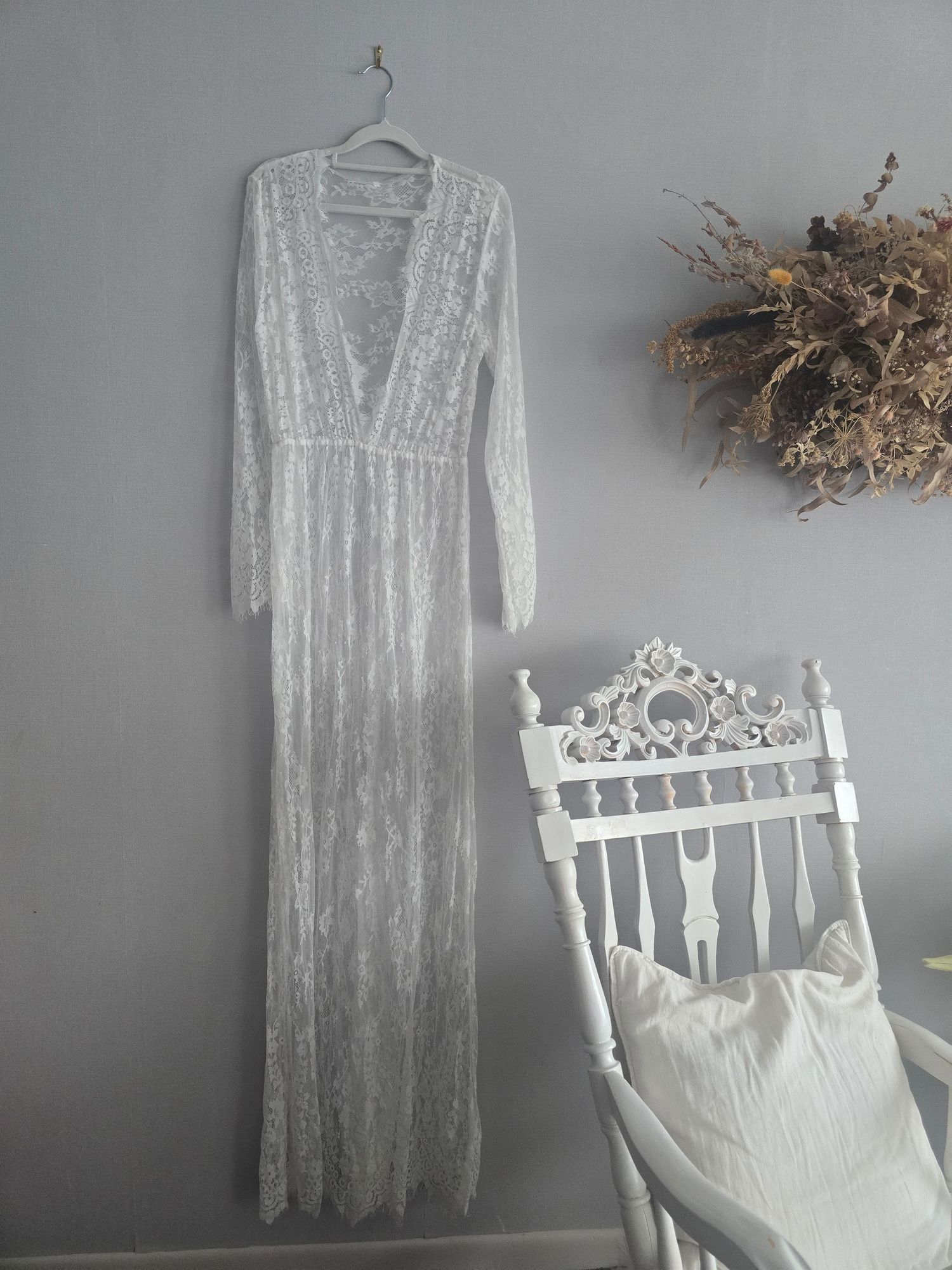 White lace long sleeve maxi dress displayed on hanger against gray wall, with ornate white chair and dried floral decor.