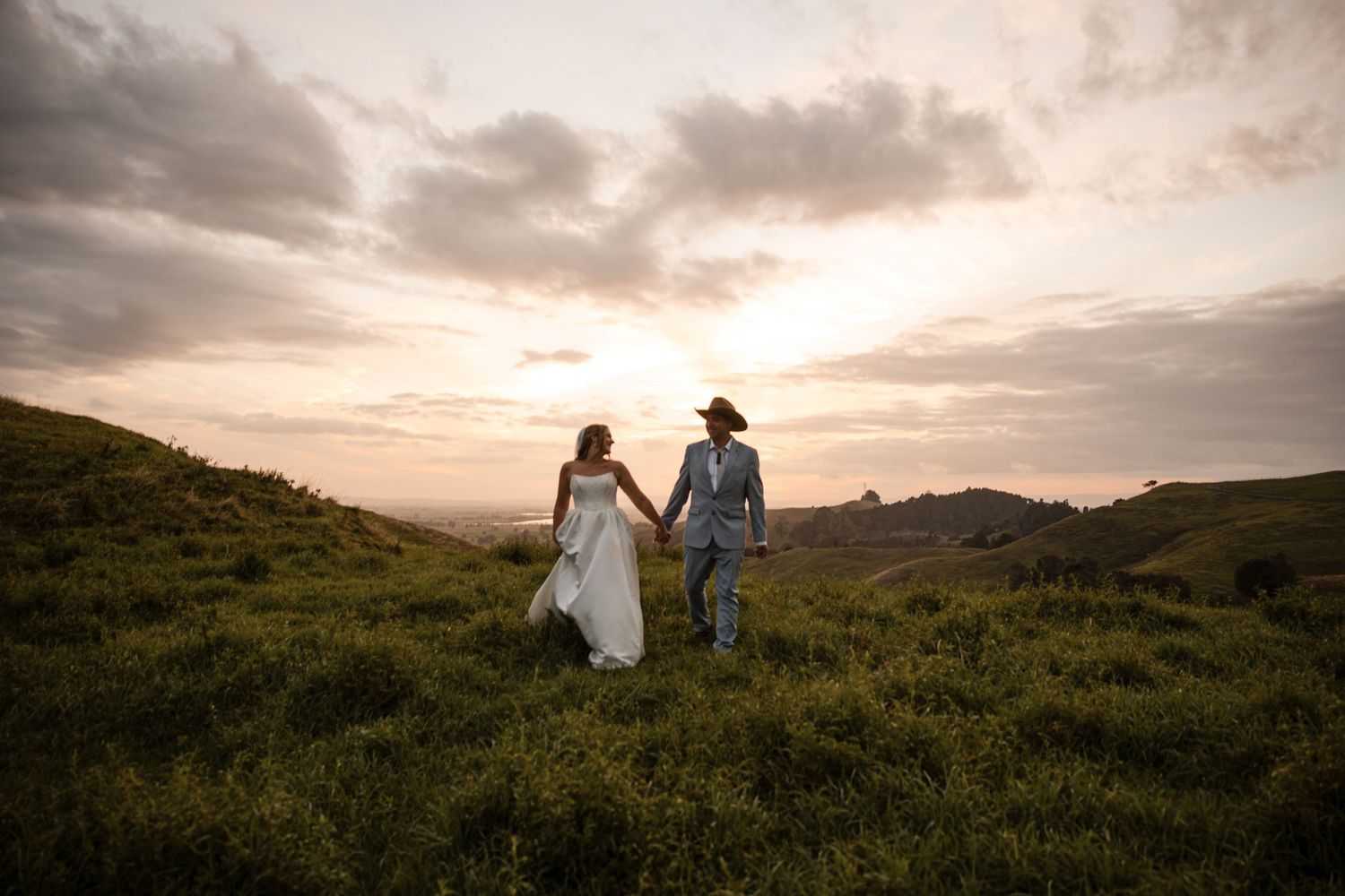 Bride and groom hold hands walking through lush green hills at sunset on their wedding day.