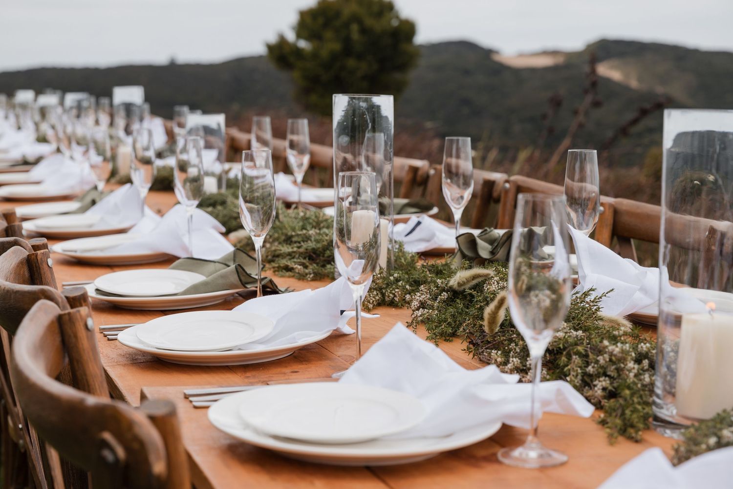 Rustic wooden dining table set with white plates, champagne glasses and greenery garland centerpiece at outdoor venue.