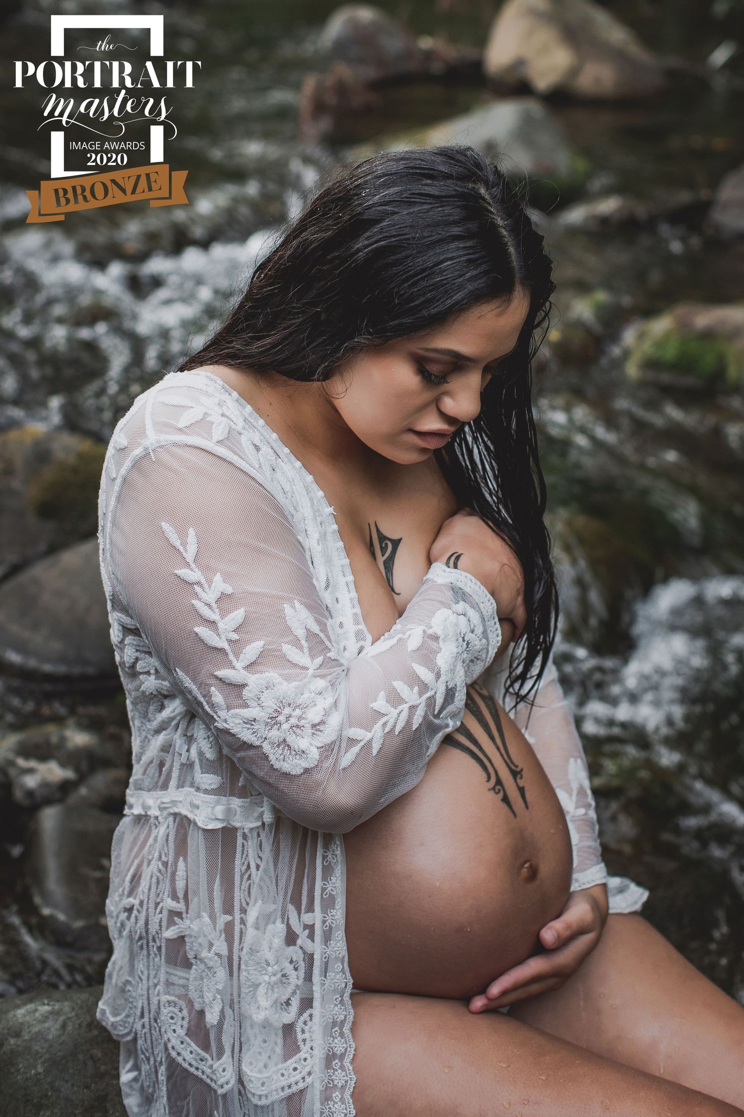 A maternity portrait shows an expectant mother in a white lace dress sitting by a stream in nature.
