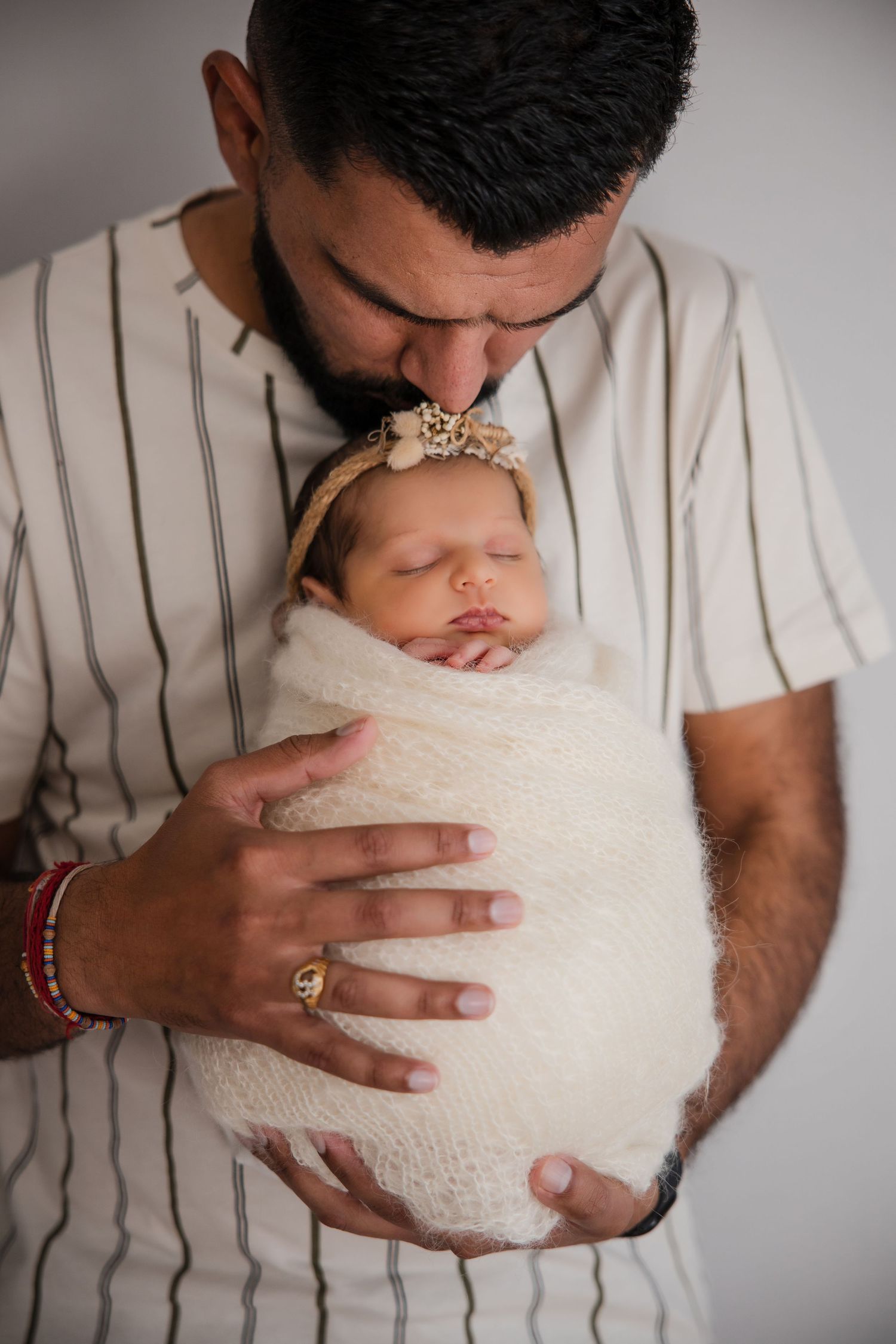 Father gently kissing swaddled newborn baby girl wearing floral headband, tender parenting moment.