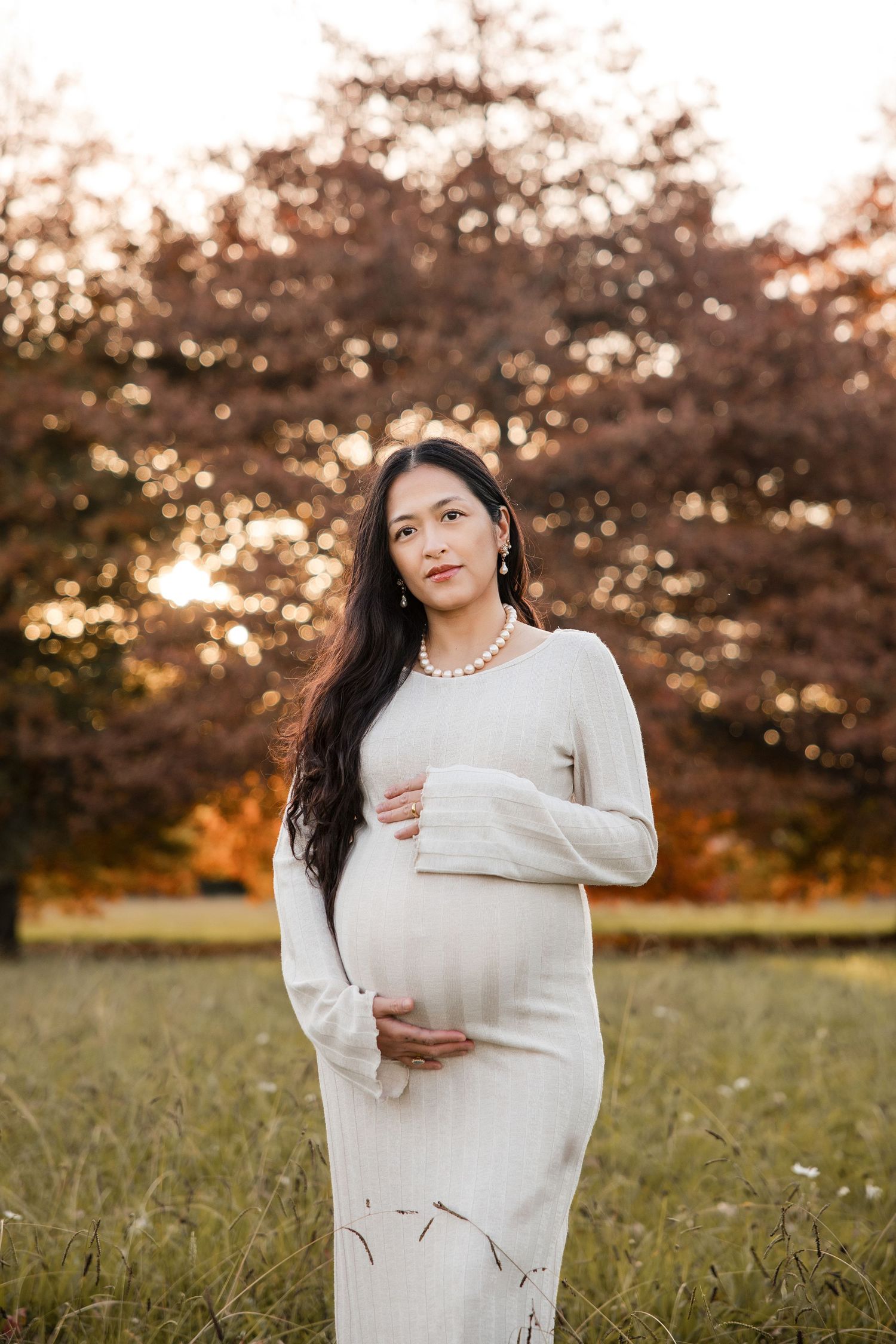 Pregnant woman in white dress cradling baby bump outdoors during golden hour with autumn trees in background.