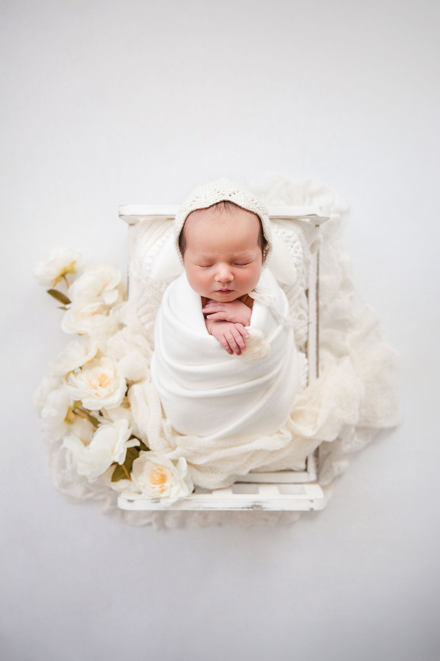 Sleeping newborn baby wrapped in white blanket on wooden tray with soft peach roses creating dreamy composition.
