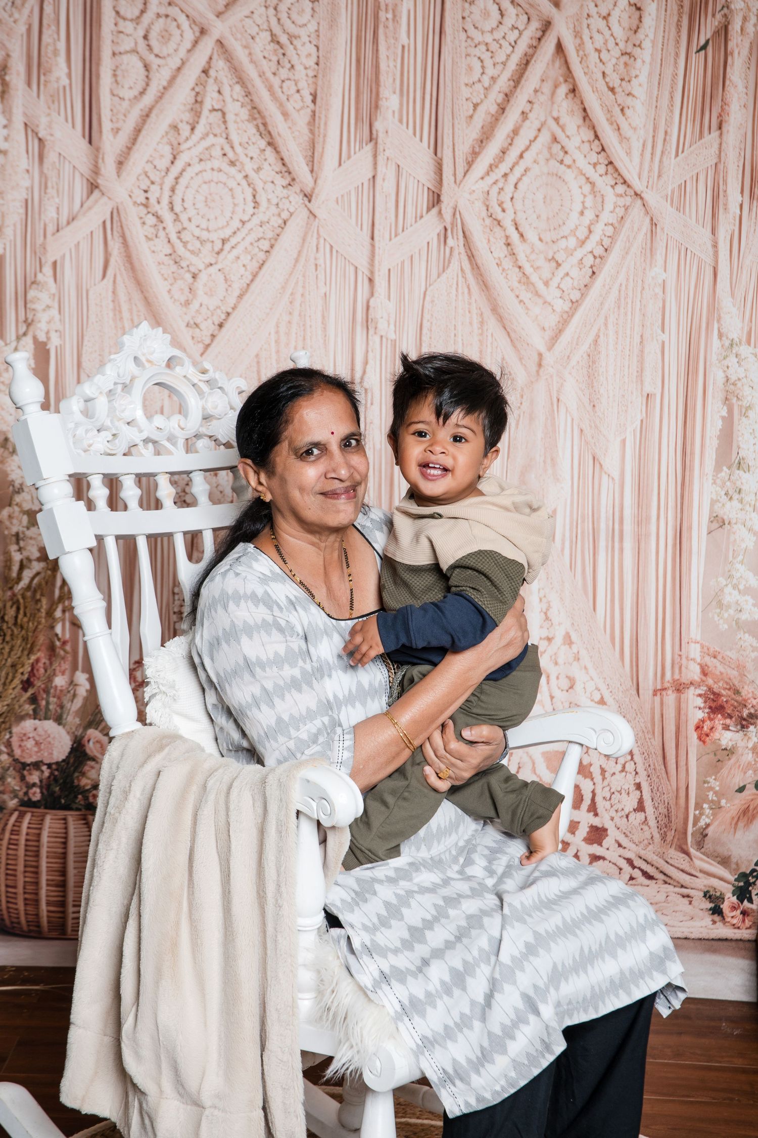 A parent and child sit together in an ornate white chair against a blush pink macrame backdrop.