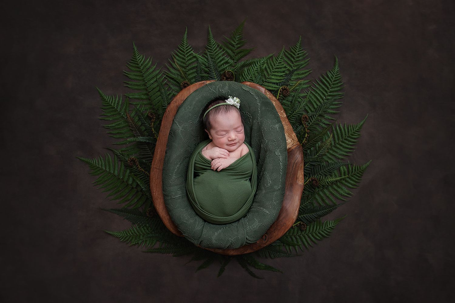Newborn rests peacefully in green wrap inside wooden bowl surrounded by fern leaves on dark background.