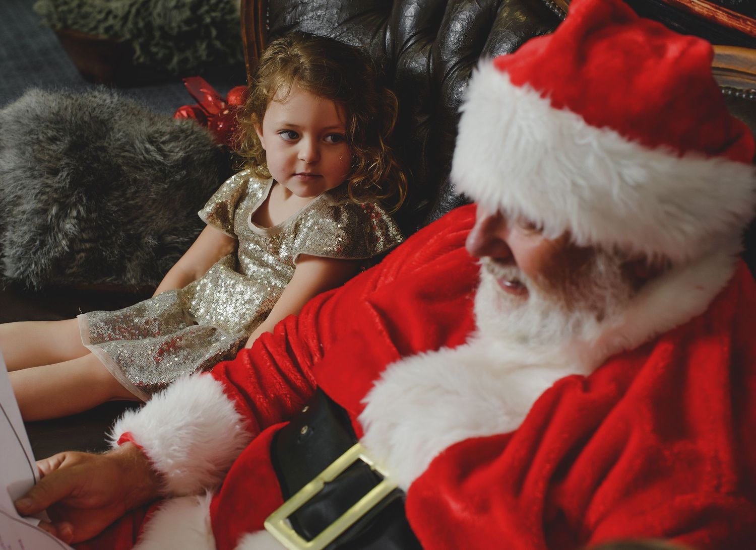 A young child in a sparkly dress sits beside Santa Claus in his traditional red suit and hat during a holiday visit.