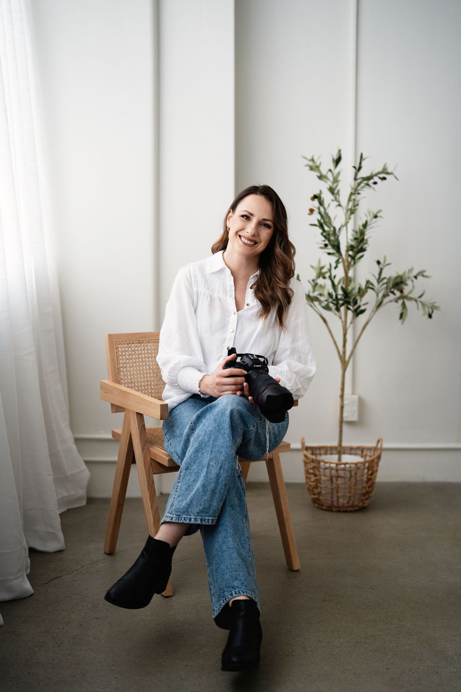Minimalist lifestyle photoshoot in white room with wooden chair, jeans, and potted plant decor.