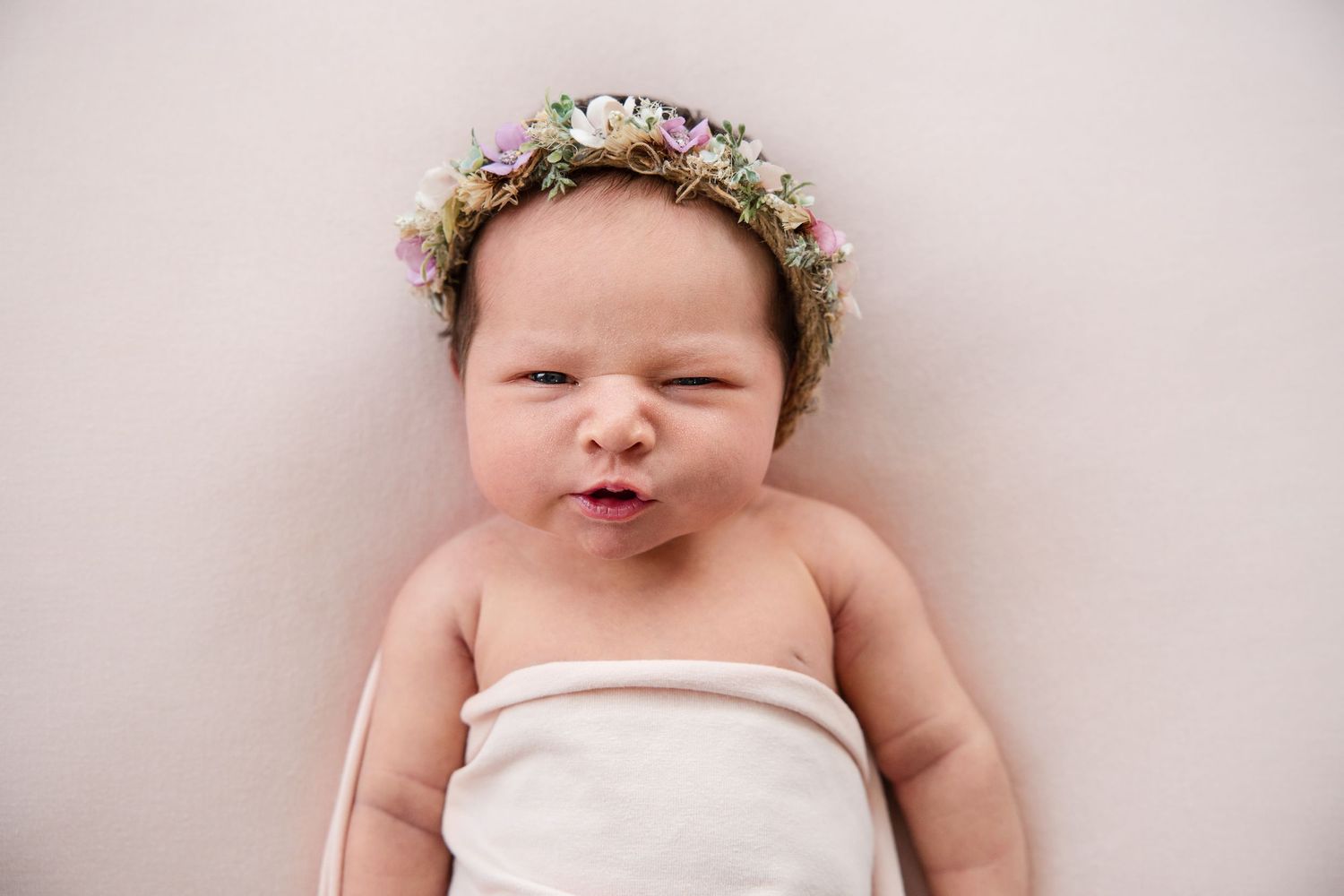 A sleeping baby wearing a floral crown and white wrap lies peacefully on a soft blanket during a newborn photo session.
