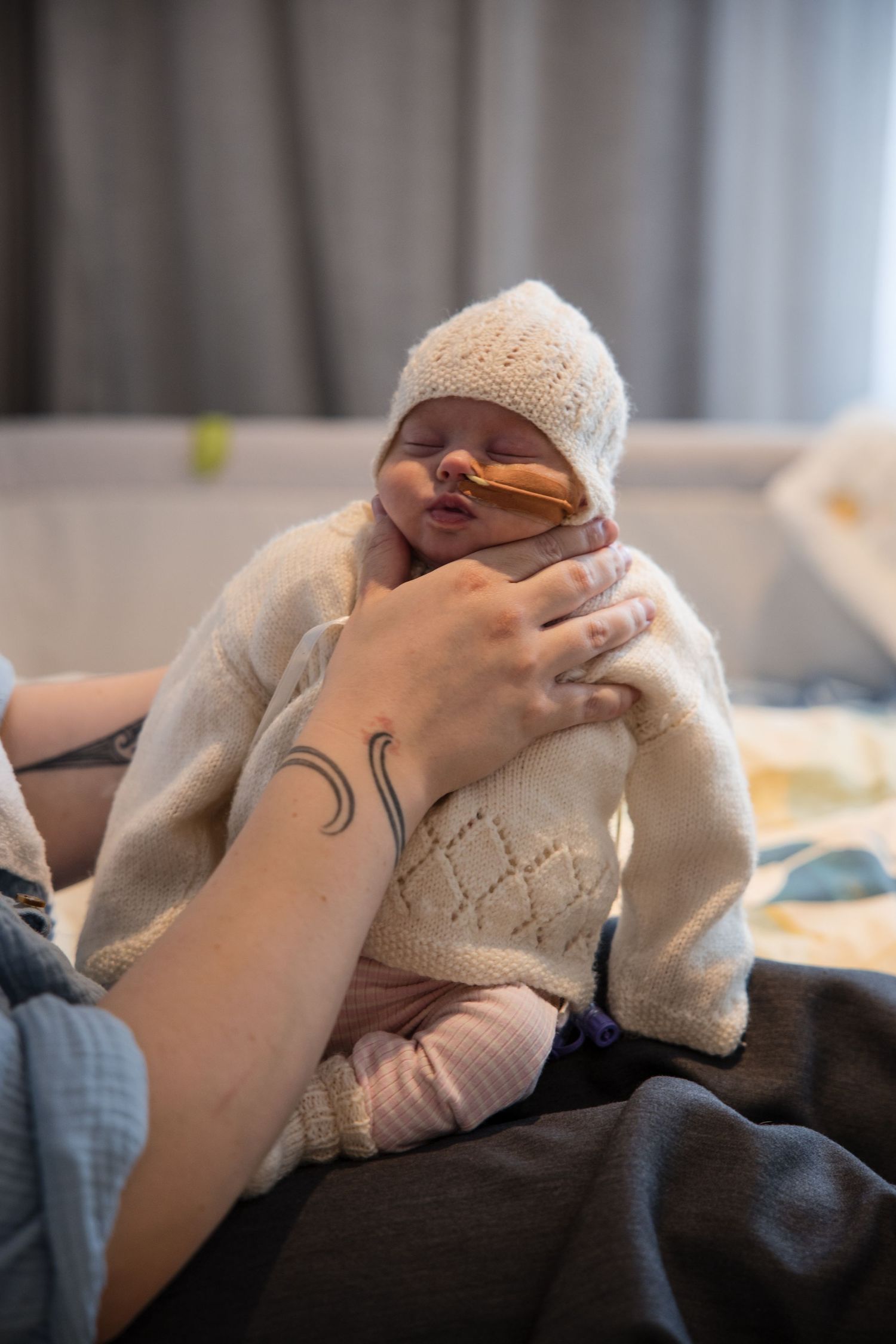 Newborn baby with feeding tube held by tattooed parent in white knit outfit and hat.