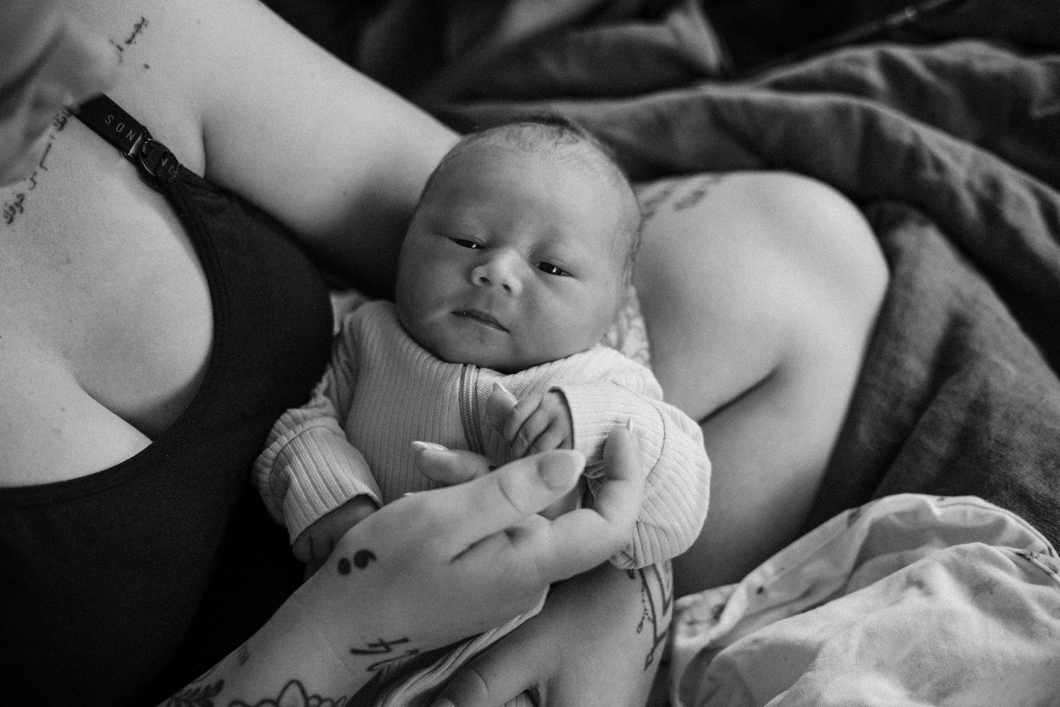 Black and white photo of a newborn baby resting peacefully in a tattooed mother's arms on a soft bed.