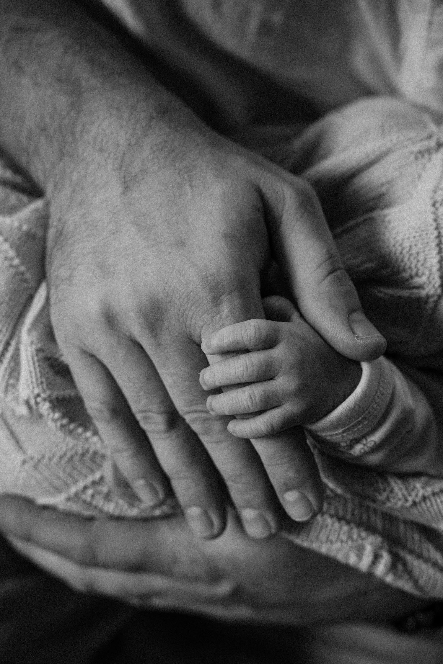 Black and white photo of a newborn baby's tiny hand grasping an adult's finger, wrapped in a soft knit blanket.