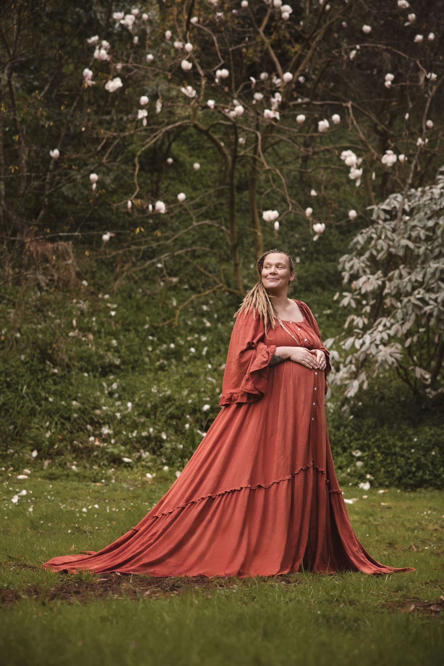 A pregnant figure in a flowing rust-colored dress stands in a garden with blooming magnolia trees in the background.