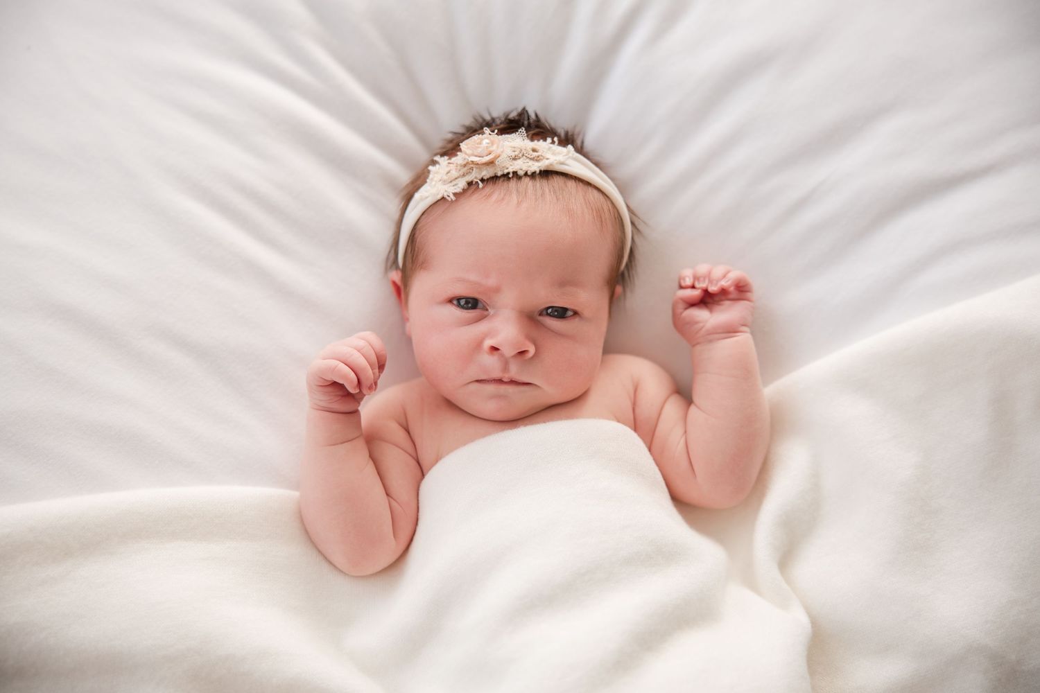 Baby in pink swaddle lying on white bedding with hands raised near face.