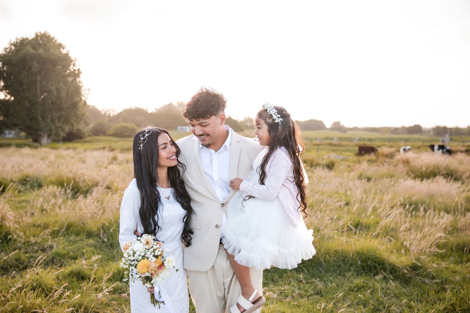 A couple in white outfits share tender moments together while walking through a golden sunlit meadow at sunset.