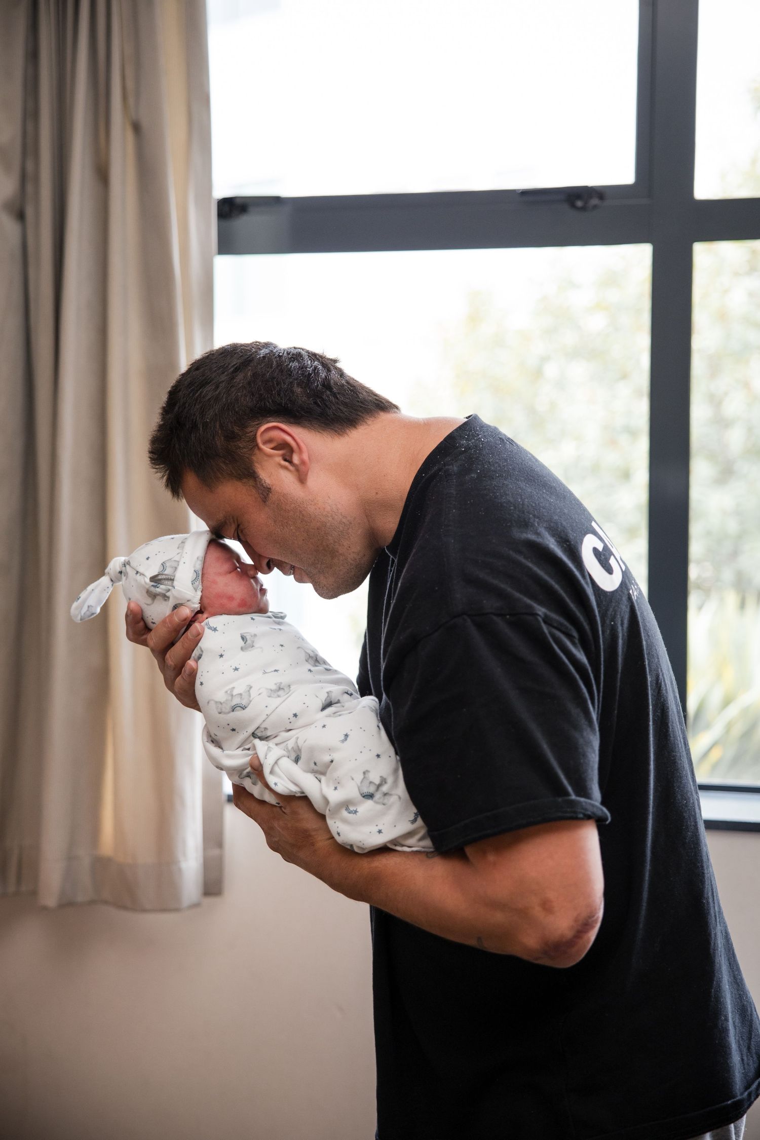 A father tenderly touches foreheads with his swaddled newborn baby near a bright window.