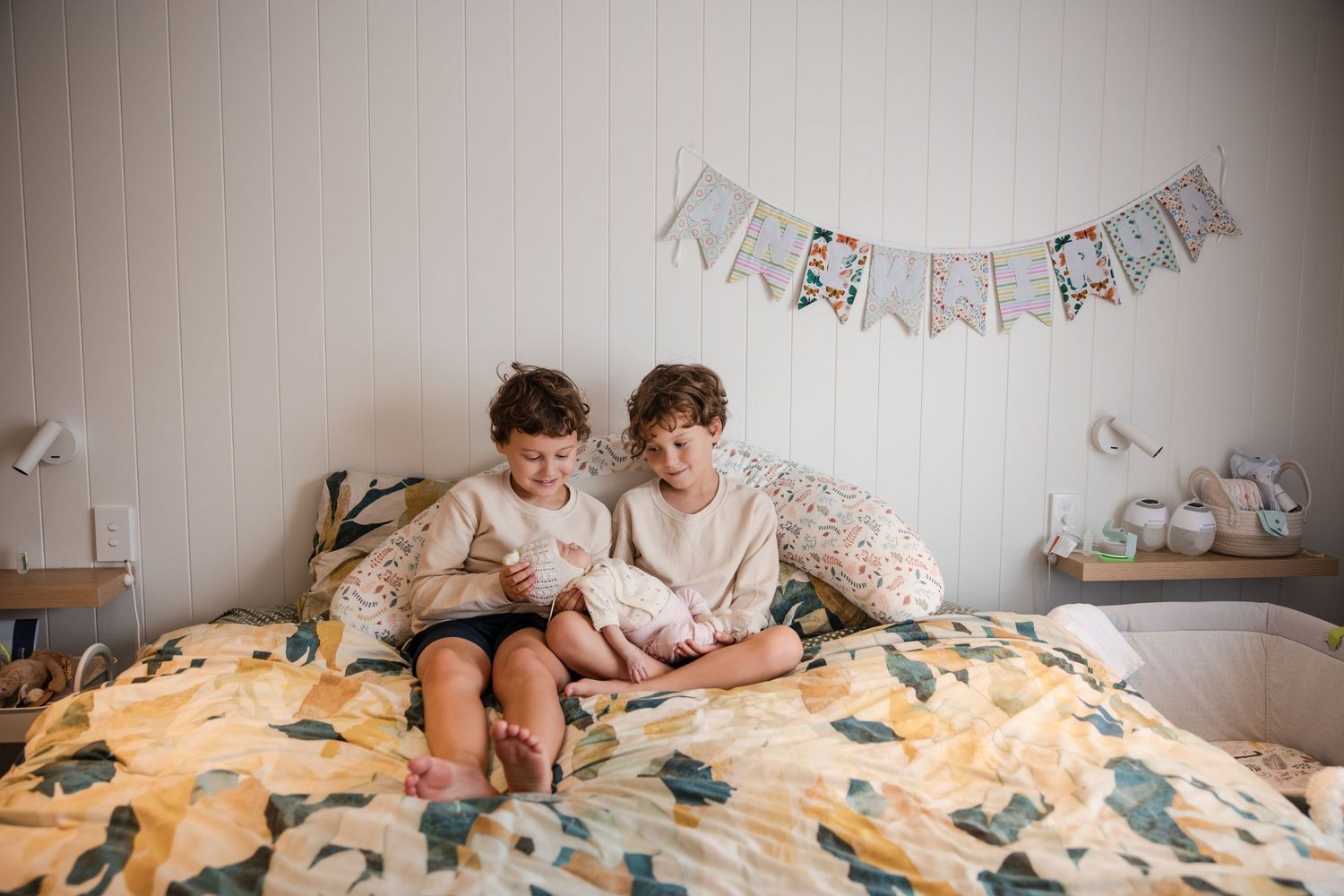 Two children sit together on a floral bedspread reading, beneath a decorative bunting banner in a cozy bedroom.