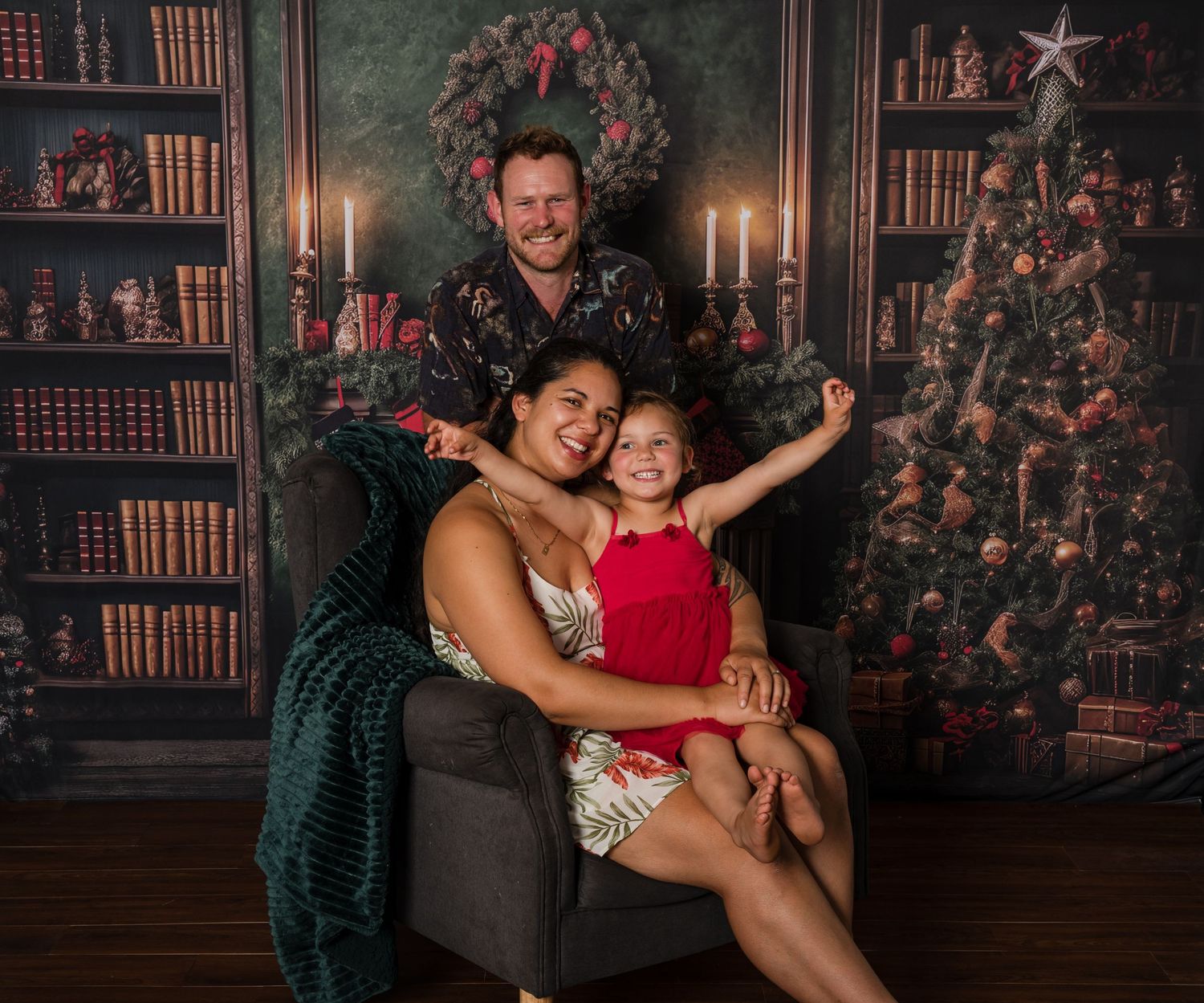Family of three poses joyfully in front of a decorated Christmas tree and bookshelf backdrop for holiday portrait.