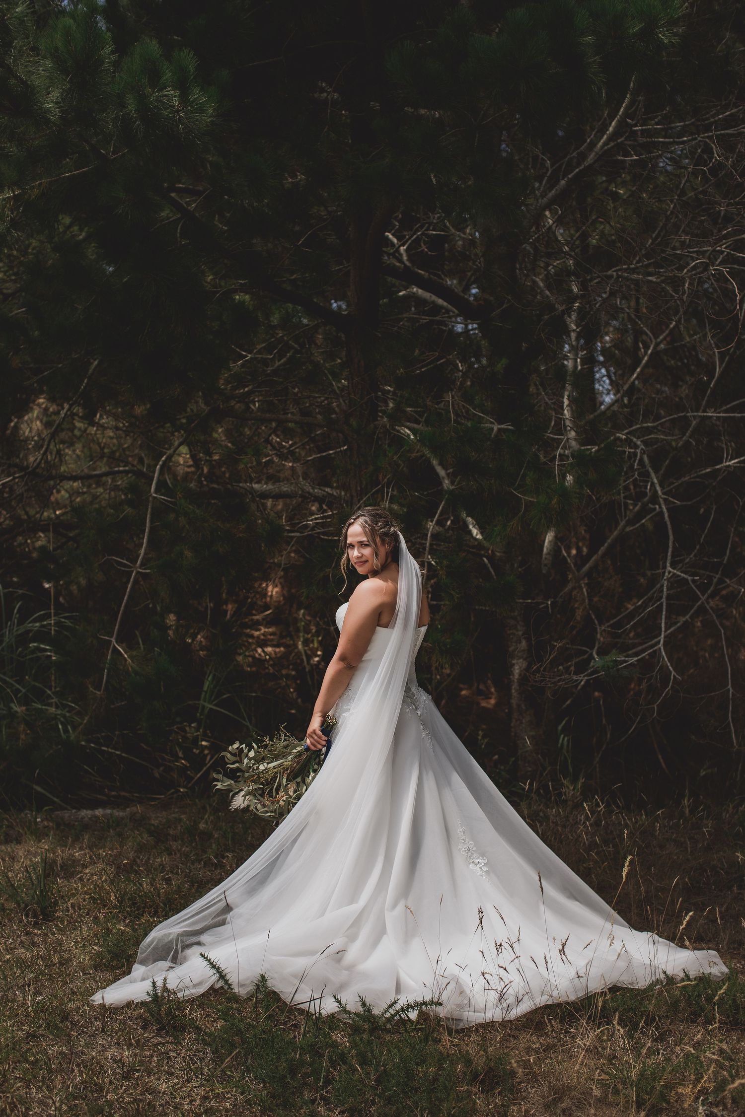 Bride in flowing white gown with train poses against dark forest backdrop. Cambridge Waikato NZ Photographer. White Locket Photography