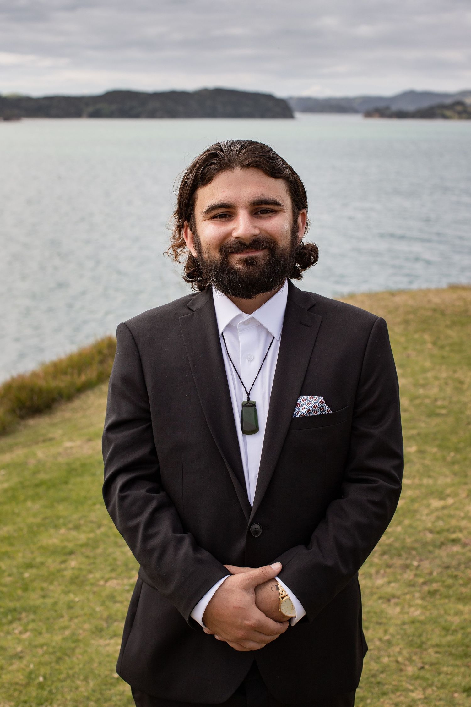 Well-dressed man in dark suit standing on grassy cliff overlooking scenic lake and mountains on cloudy day.