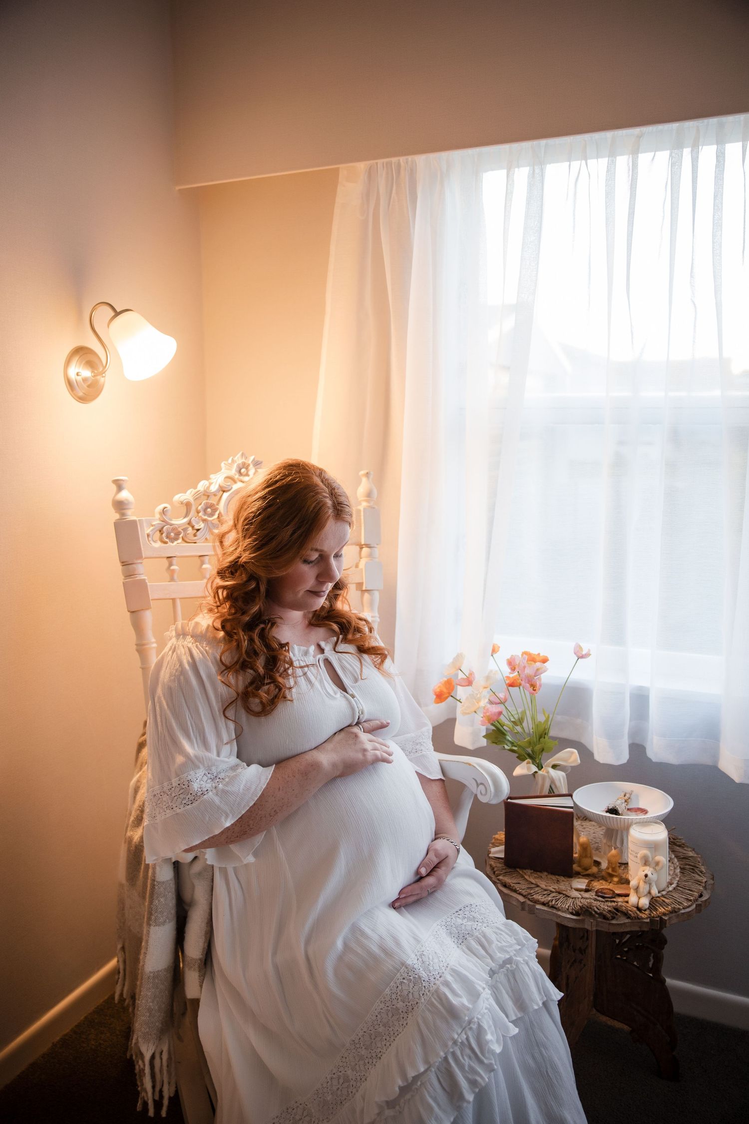 A pregnant woman in a white lace dress sits in an antique chair by a window with soft lighting and floral decor.