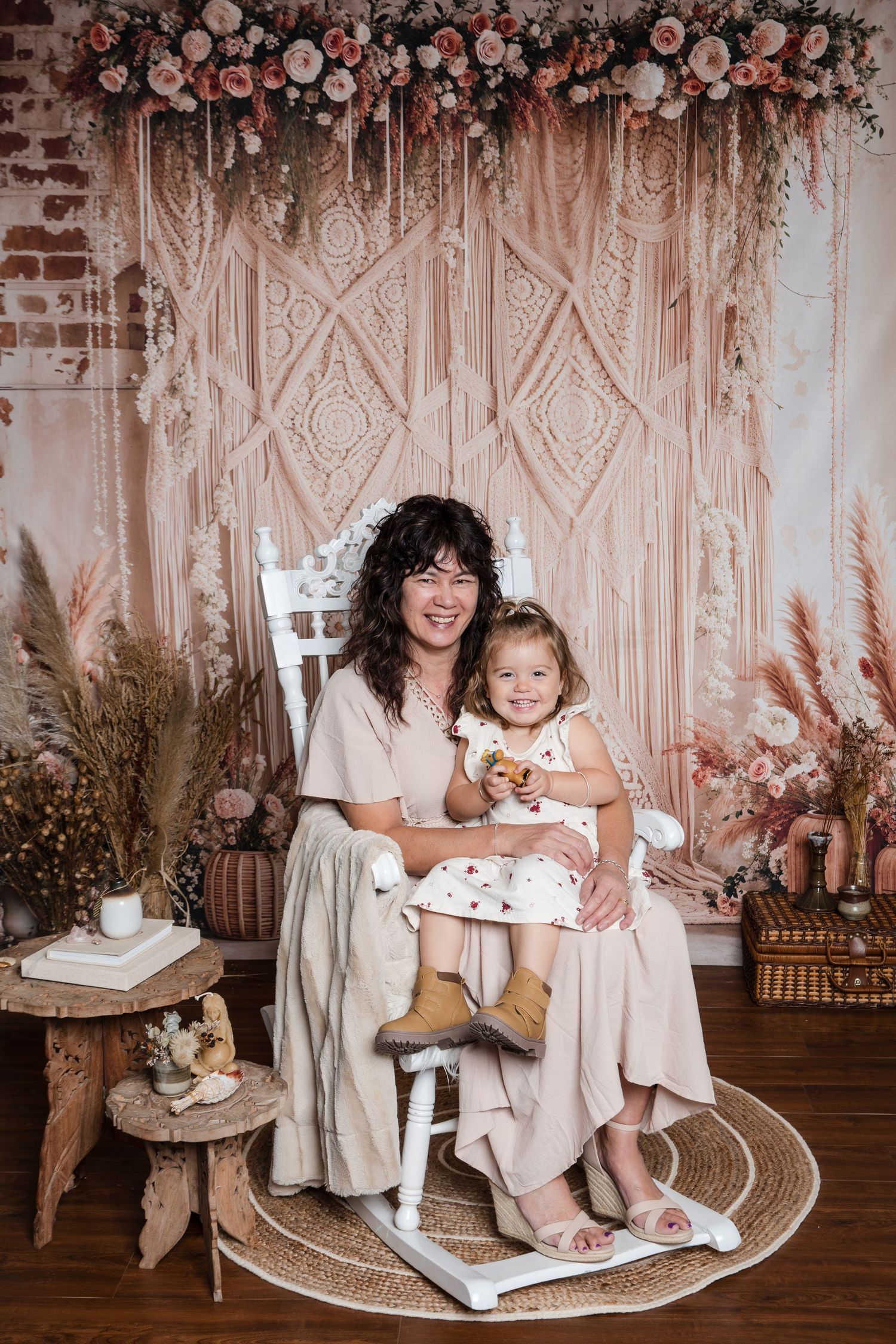 Boho-styled photo session featuring macramé backdrop, rattan chair, and soft pink floral arrangements in romantic setting.