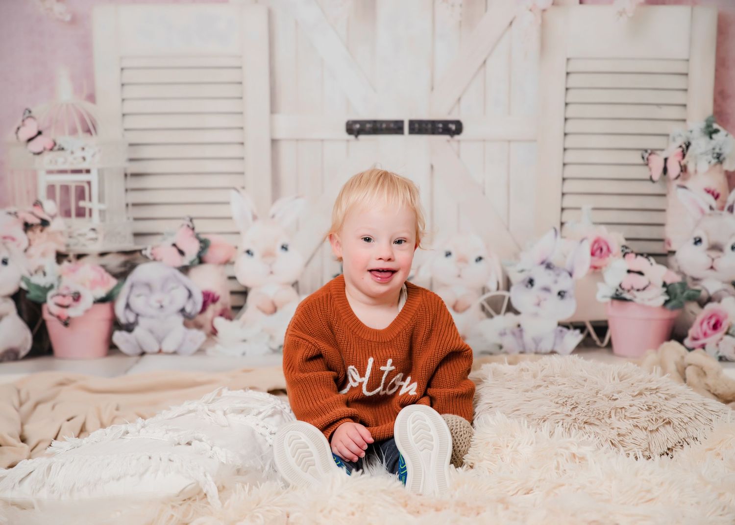 A child in a rust-colored sweater sits on cream bedding with pink floral decorations and white shutters in the background.