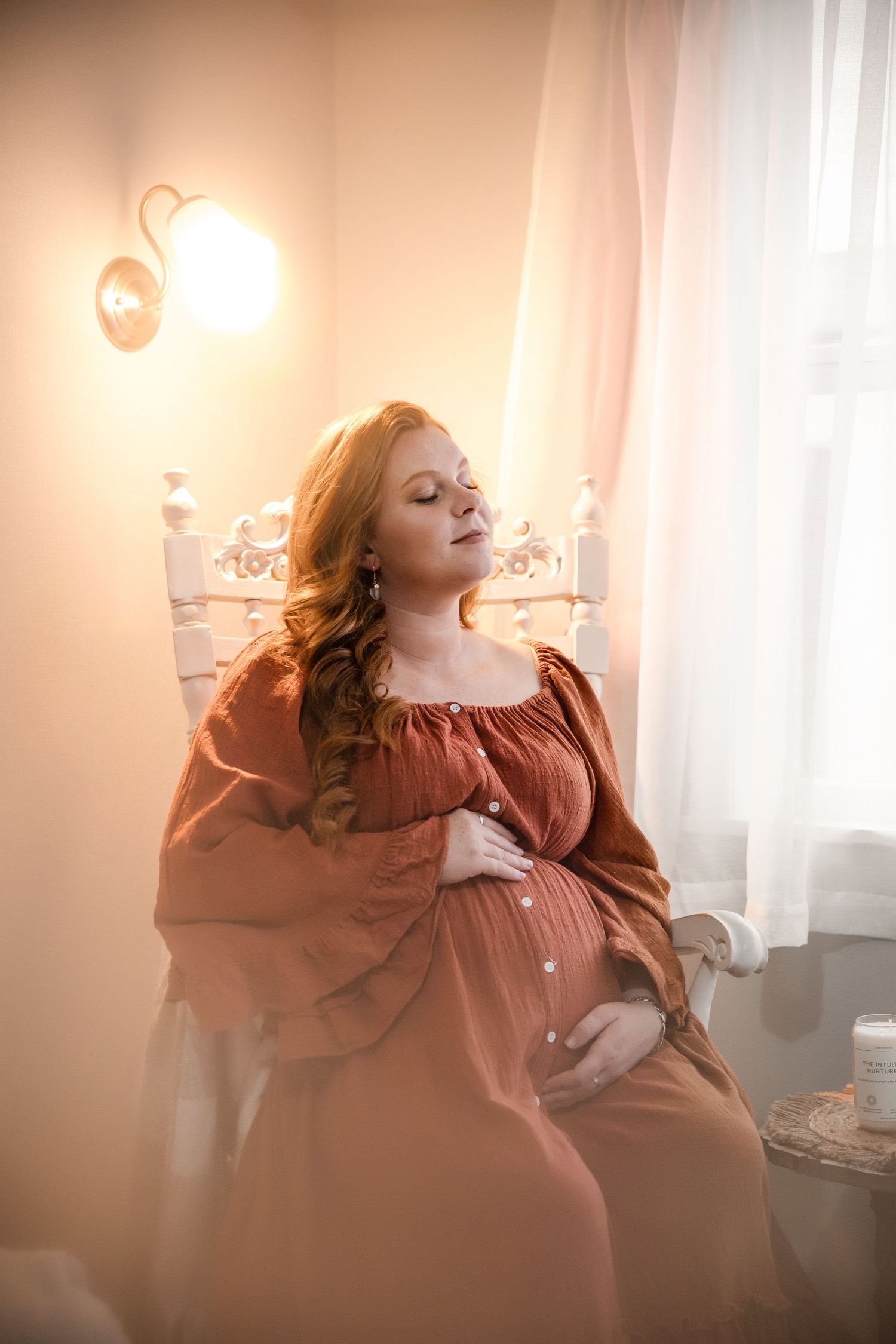A pregnant woman in a rust-colored dress poses elegantly against a soft, window-lit interior setting.