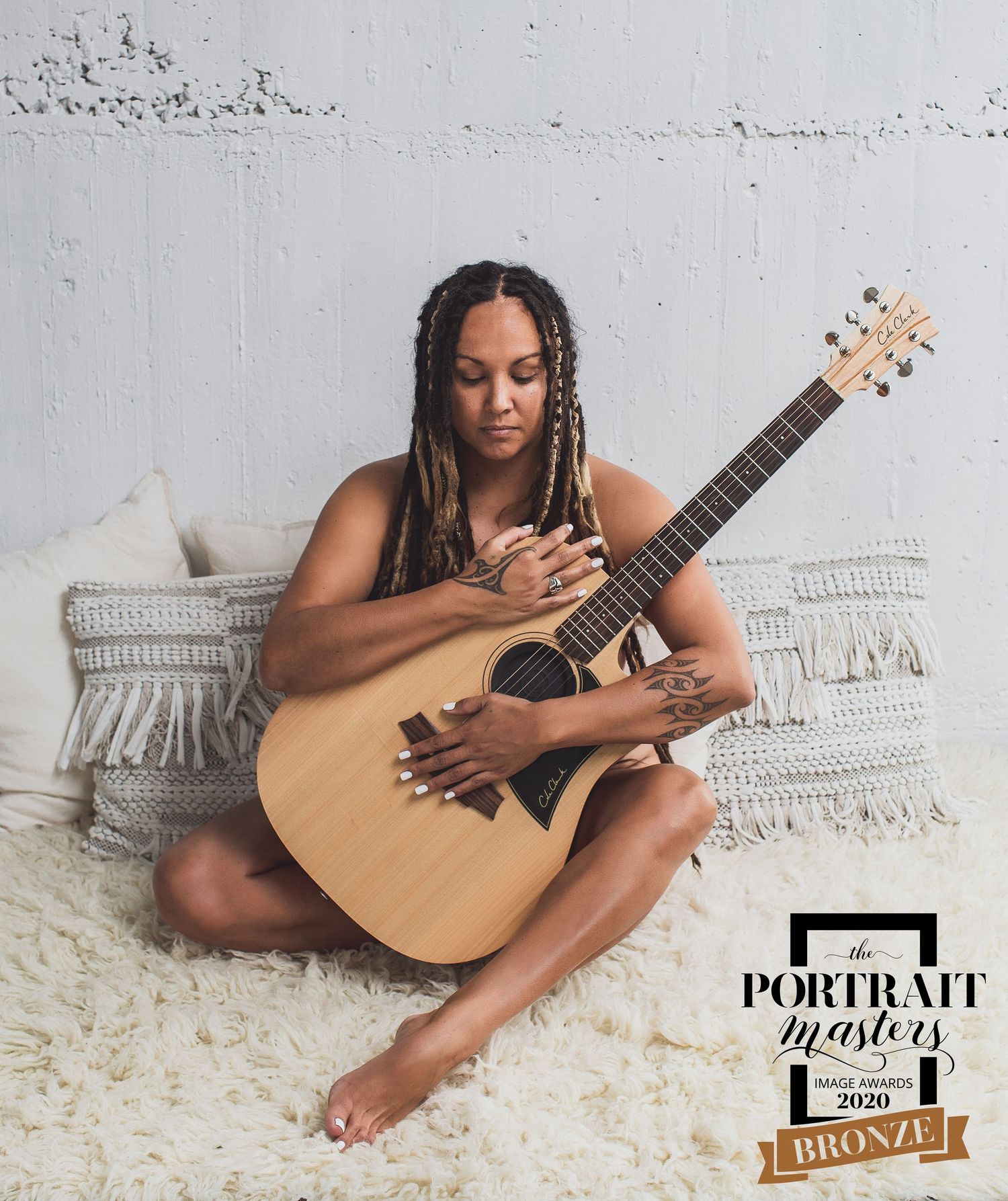 A guitarist with braided hair sits cross-legged on the floor, holding an acoustic guitar against a light colored background.