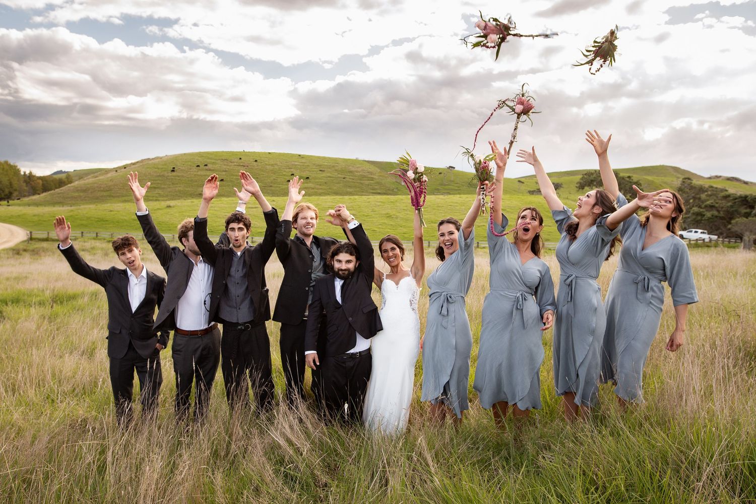 A wedding party jumps joyfully in a grassy meadow with blue sky and rolling hills in the background.