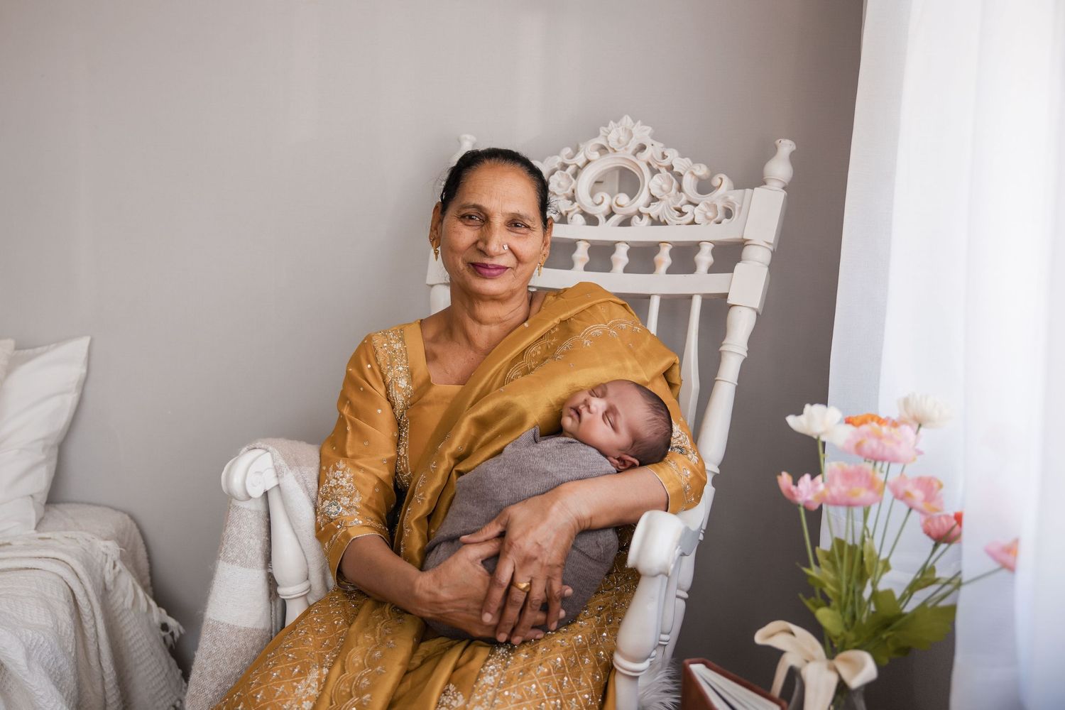 A person wearing a golden dress sits in an ornate white chair while holding a sleeping baby in a light-colored nursery setting.