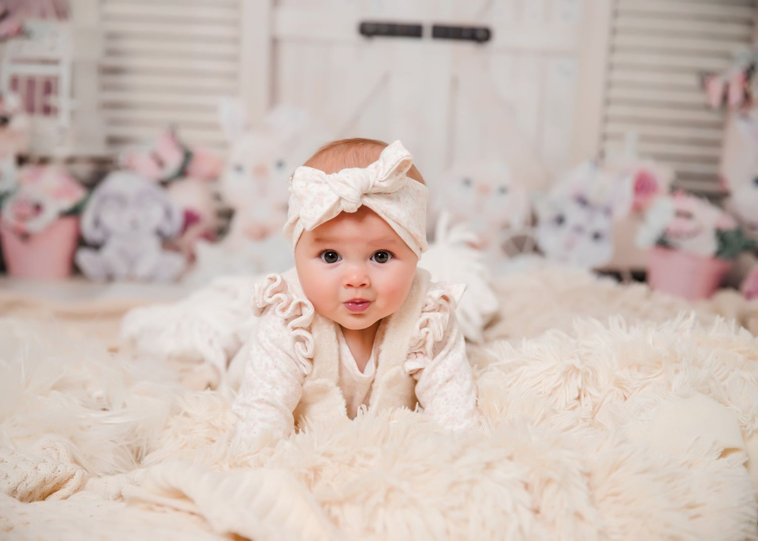 A baby wearing a cream-colored headband lies on a fluffy white blanket with stuffed animals in the background.