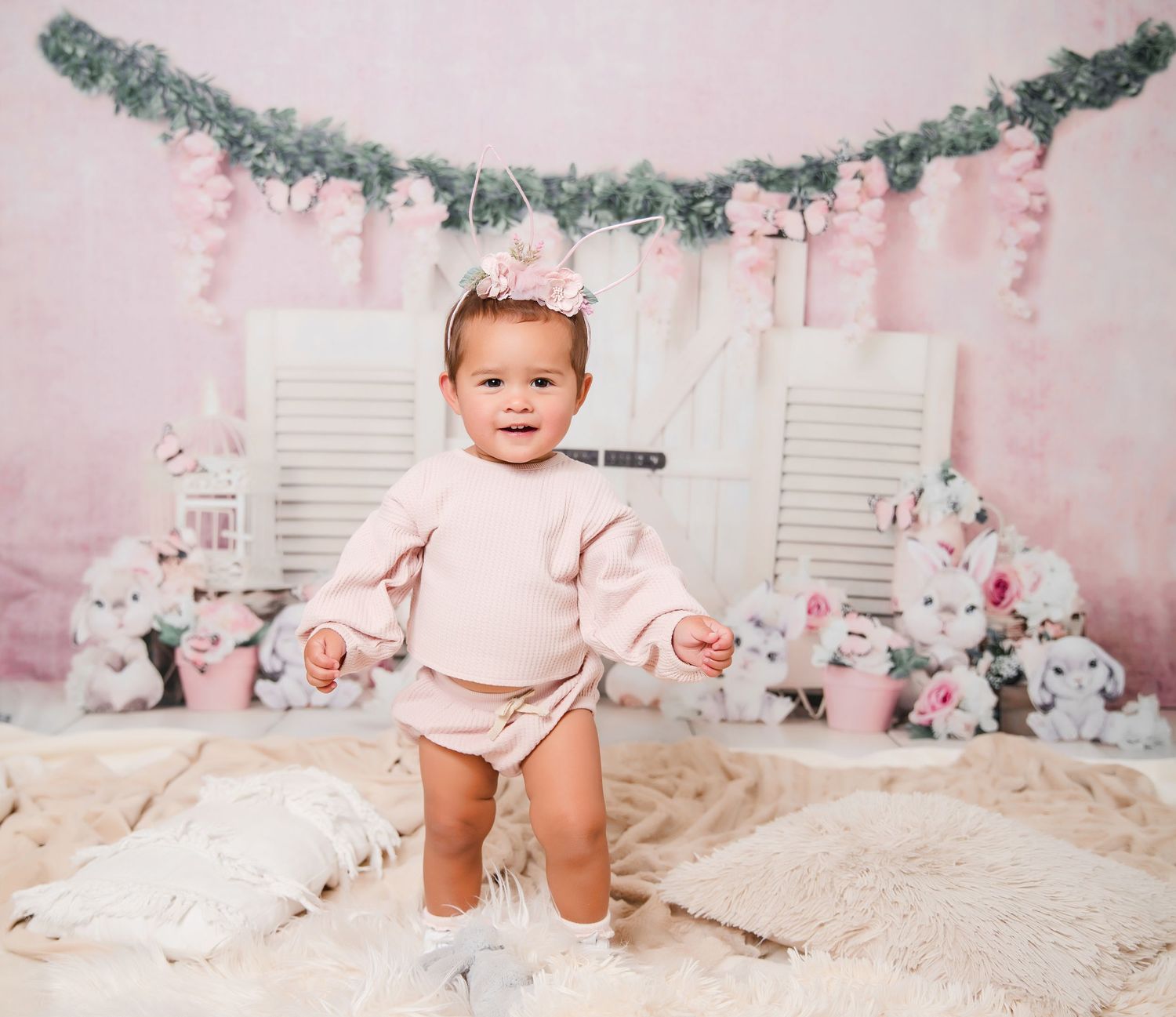 Pink and white holiday photo setup with greenery garland, white shutters, and plush toys in pastel nursery setting.