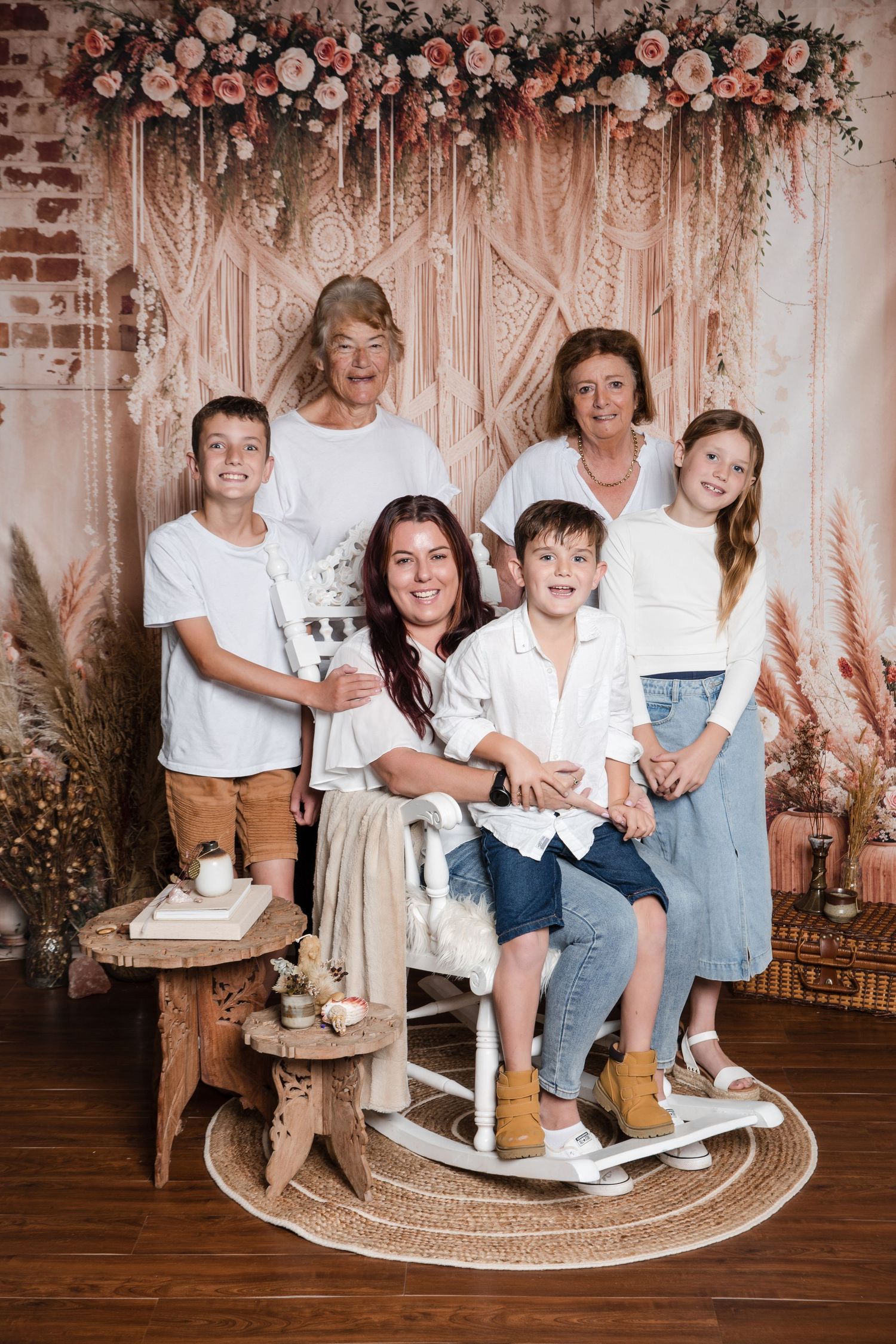 A family posed together in front of a floral backdrop with draped fabric and soft pink lighting on hardwood floors.
