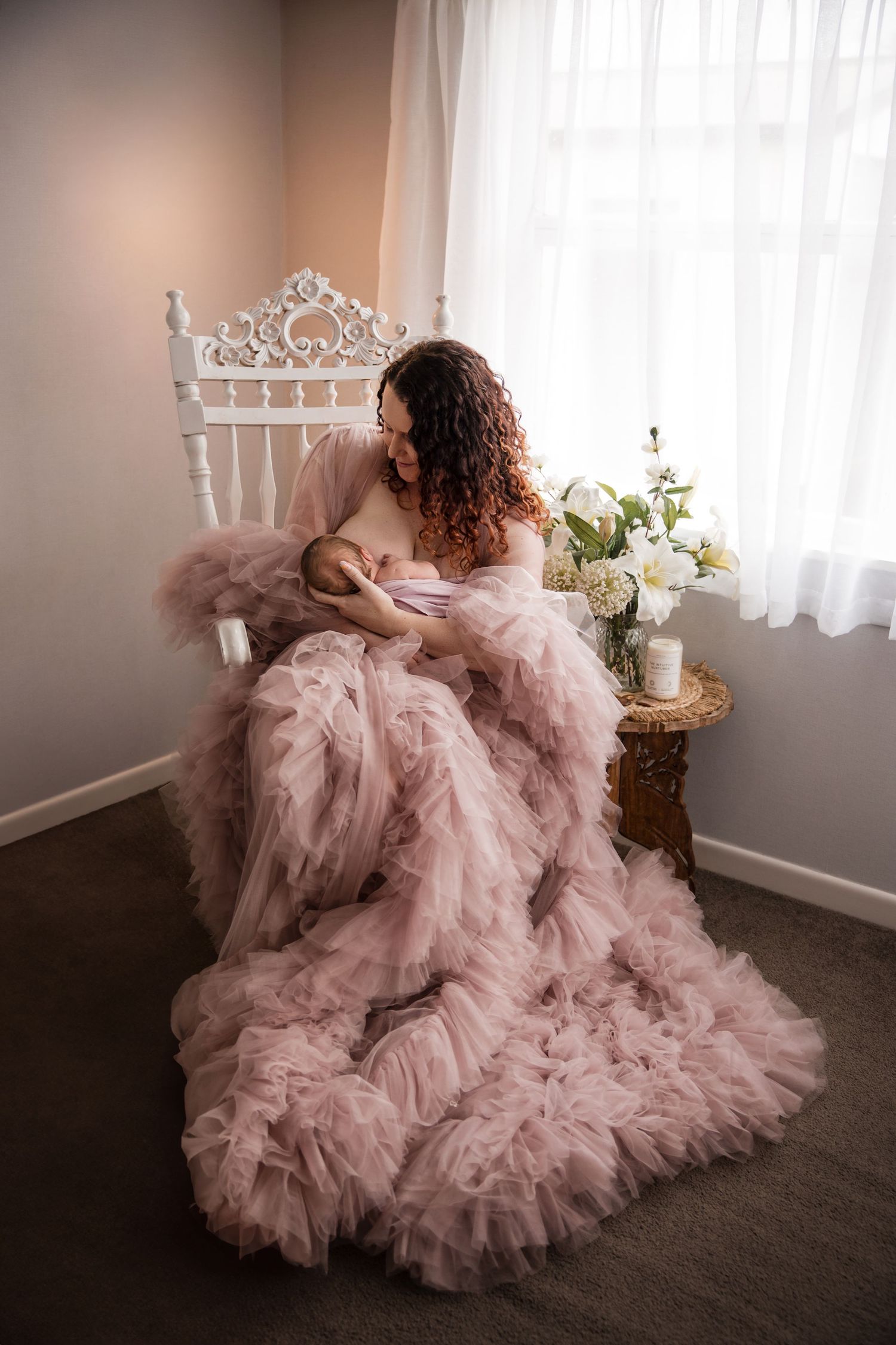 A mother in a flowing pink tulle gown sits in a white ornate chair, cradling her newborn baby near a bright window.