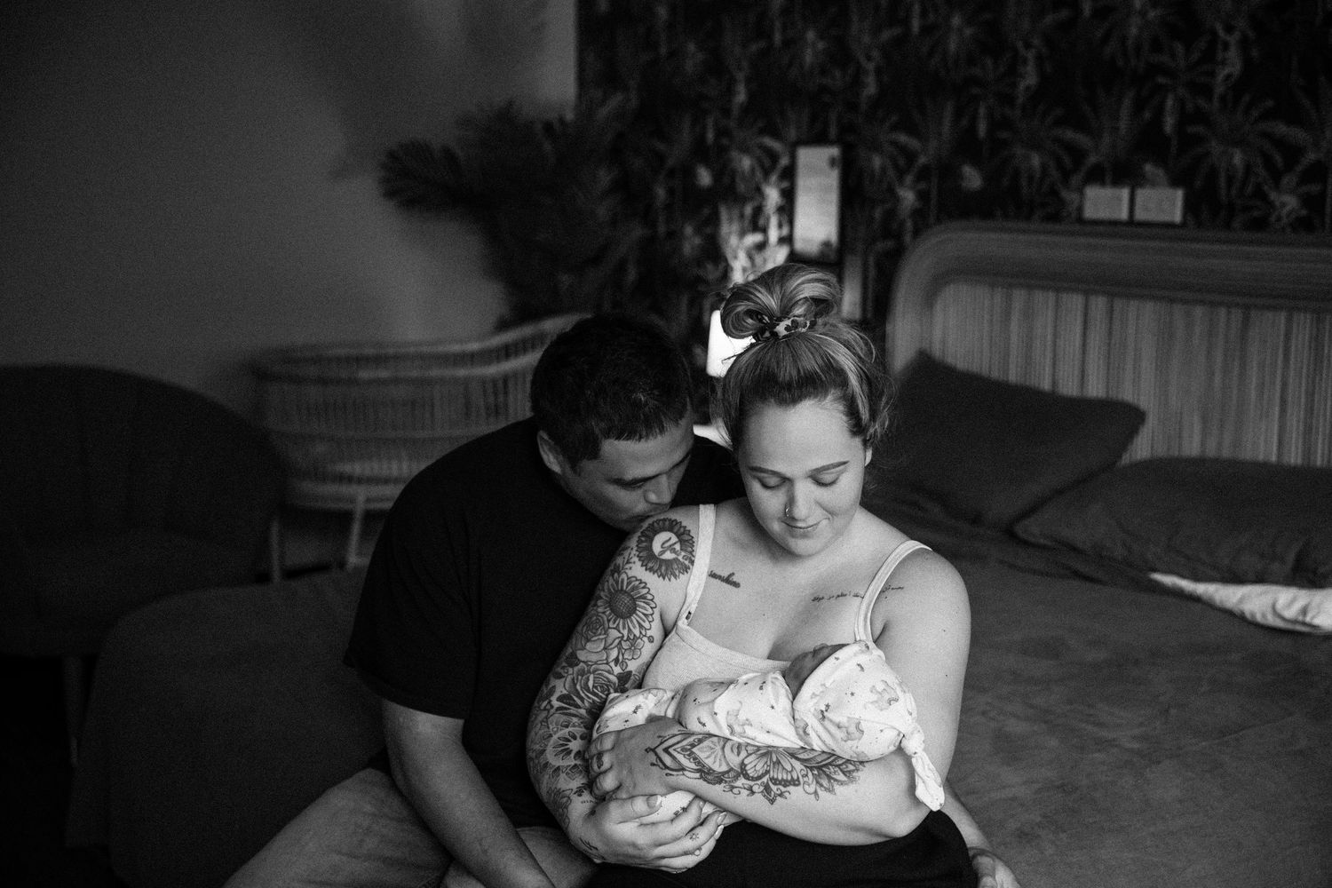 Black and white photo of a couple sitting on a bed, lovingly cradling their newborn baby together.