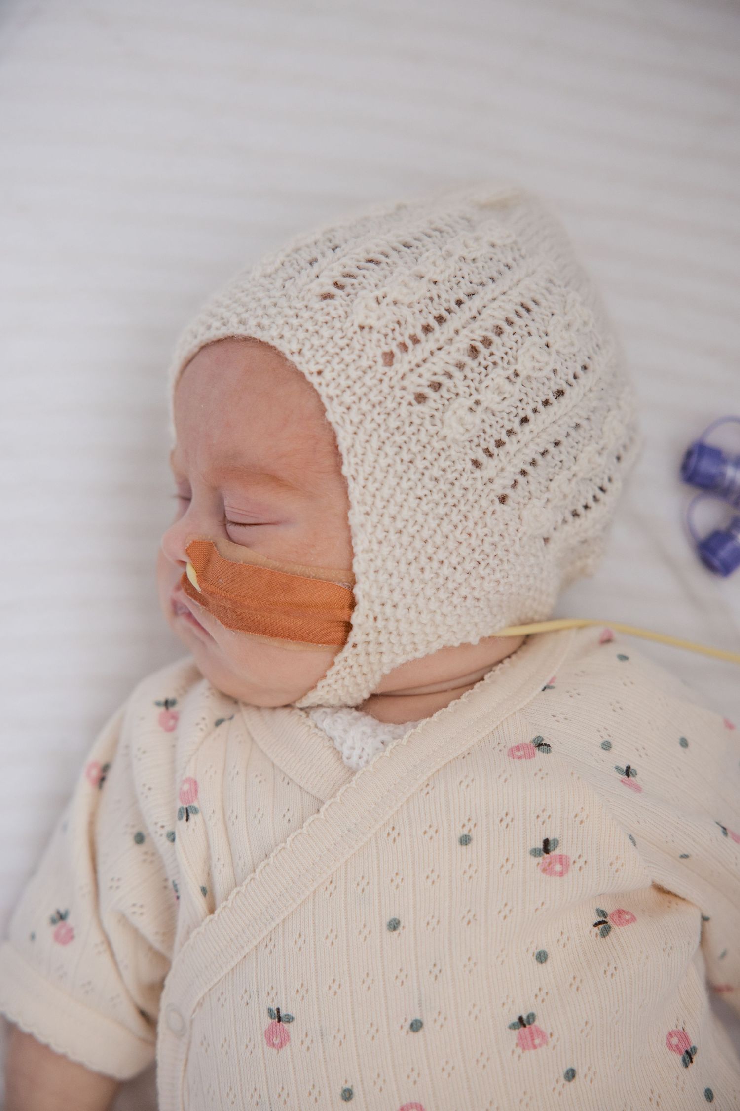 Sleeping newborn in cream knit bonnet and floral onesie with nasal feeding tube.