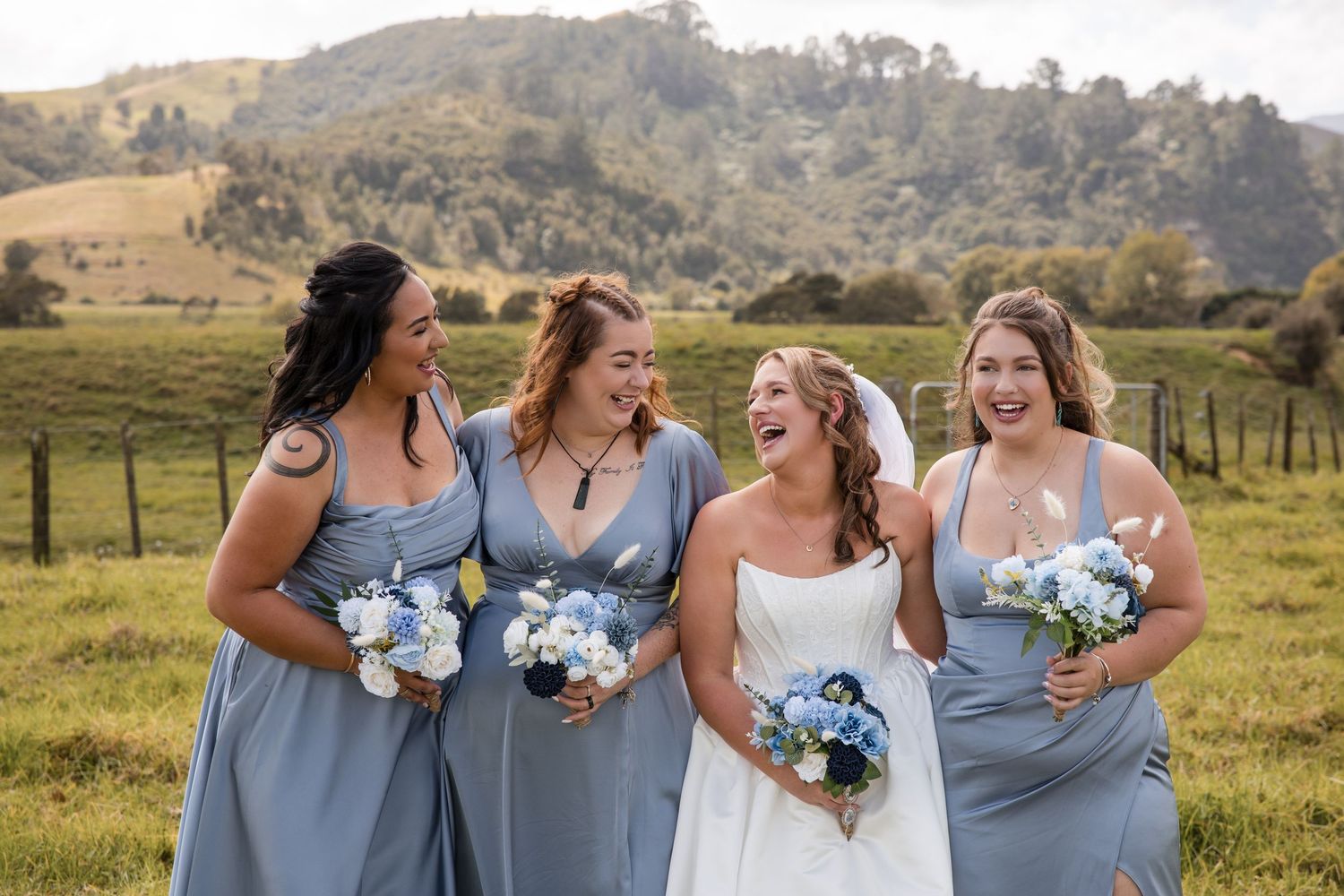 Bride and three bridesmaids in blue dresses laughing outdoors with bouquets, rolling green hills in background.