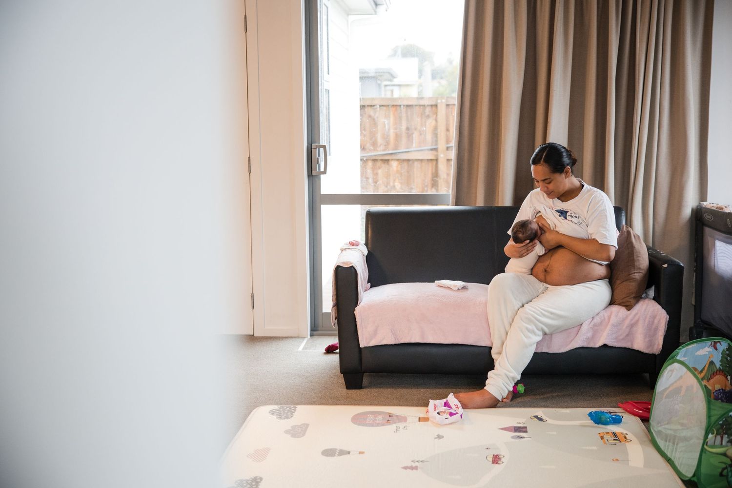 Pregnant woman sitting on a bed in a cozy room, cradling her baby bump near a large window.