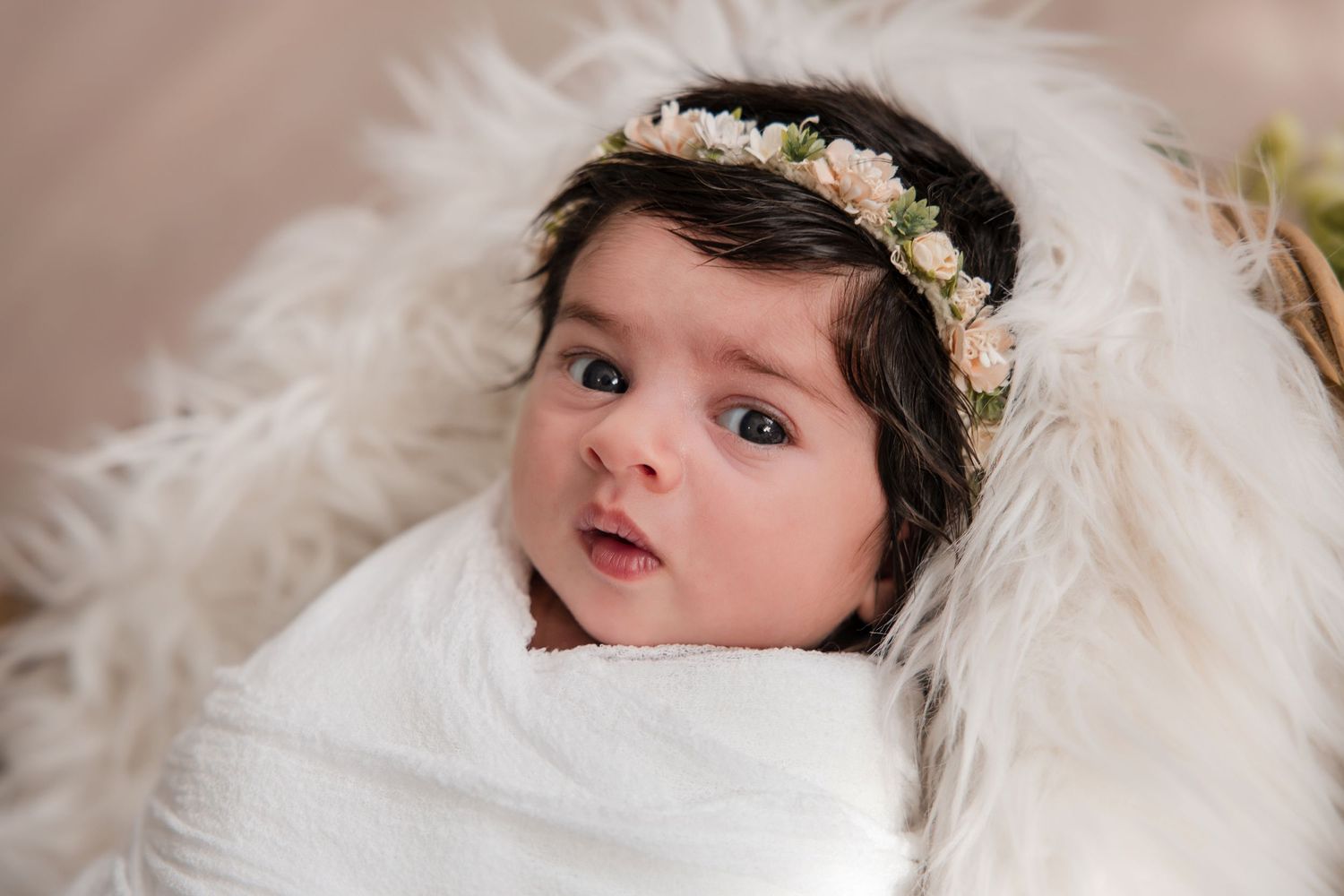 Newborn baby wrapped in white swaddle with floral crown, lying on fluffy white fur, wide-eyed and alert.