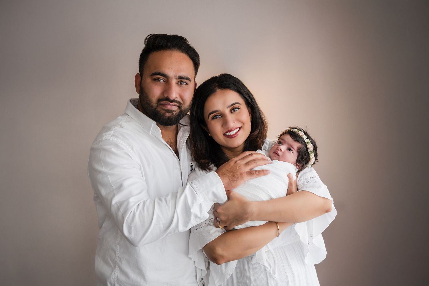 A couple in white outfits smiling while holding their newborn baby dressed in white against a neutral background.