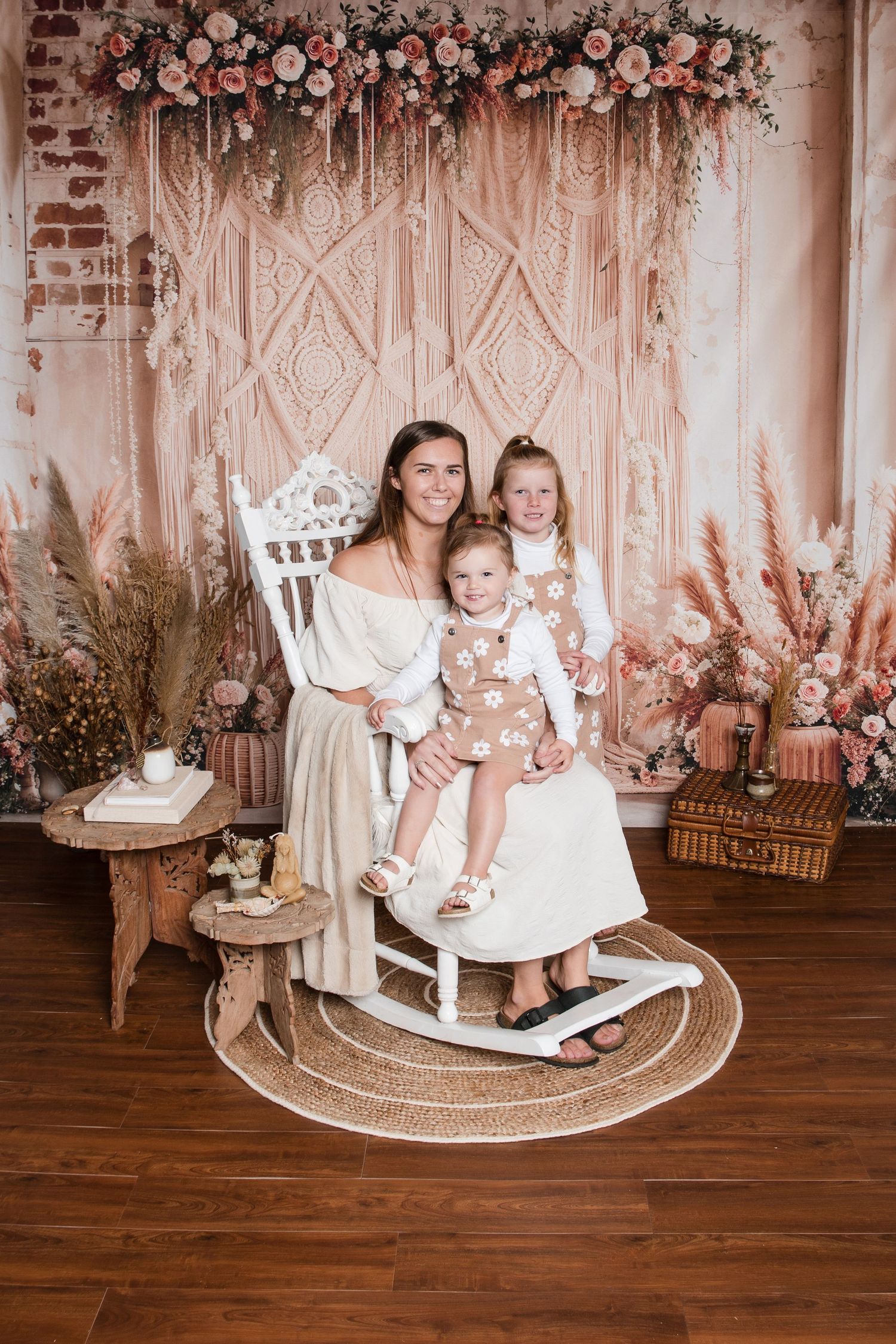 Woman and two young girls pose in a rocking chair with boho floral backdrop in a professional photo studio.