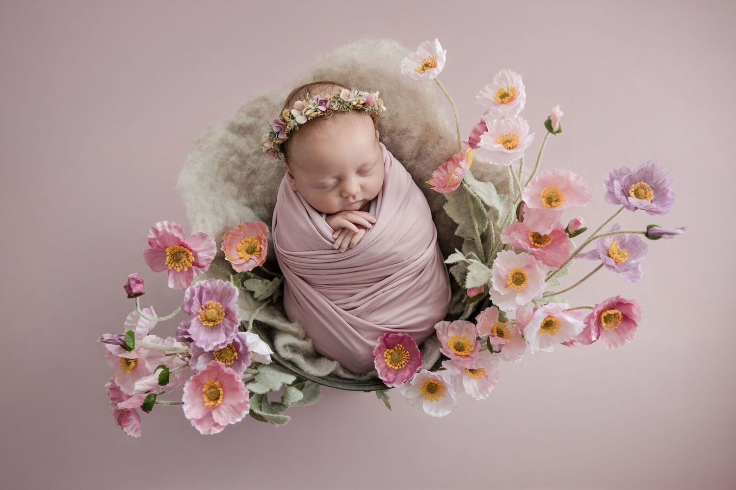Sleeping newborn baby girl wrapped in pink, wearing a floral headband, nestled in a basket surrounded by pink flowers.