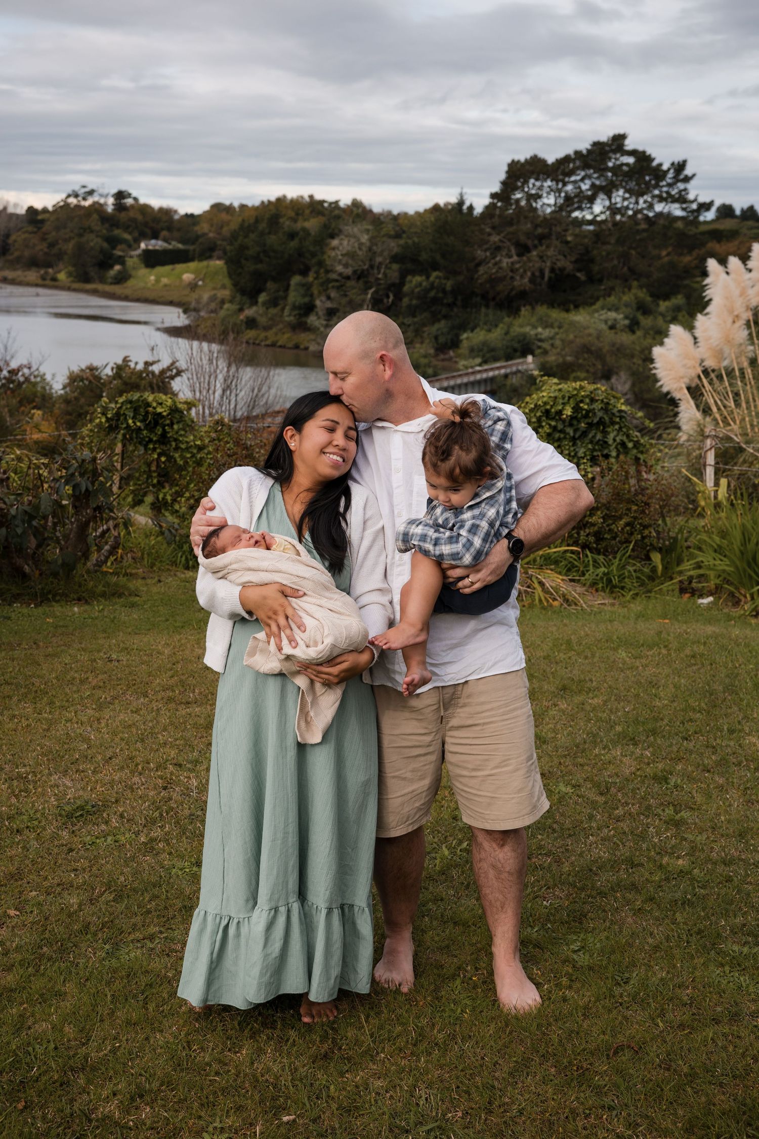A happy family of four poses outdoors, parents holding two young children with a scenic waterway and lush greenery behind them.