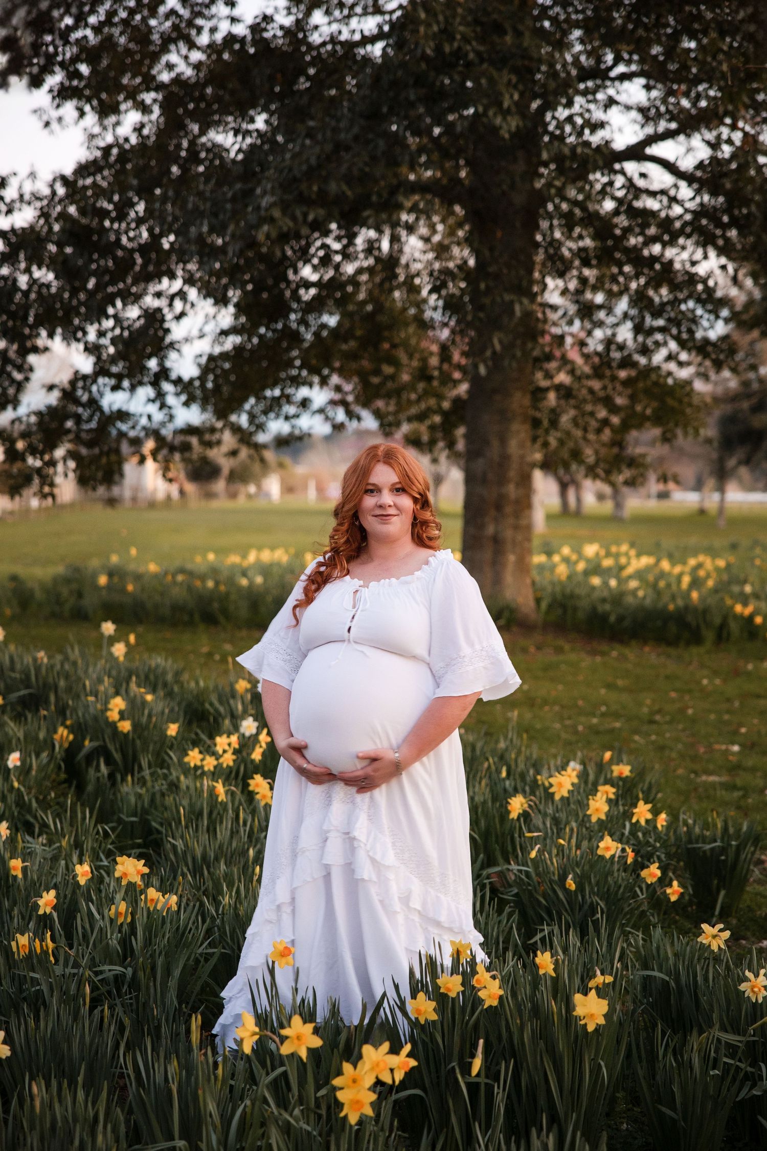 Pregnant woman in white dress holding belly among yellow daffodils in a lush green park setting.