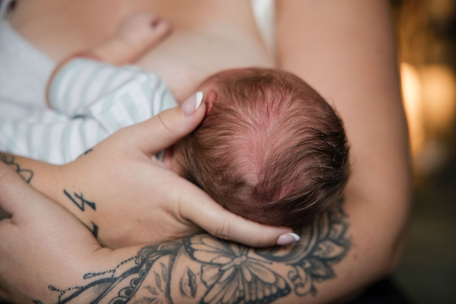Mother with floral tattoo breastfeeding her newborn baby with red hair in a tender, intimate moment.