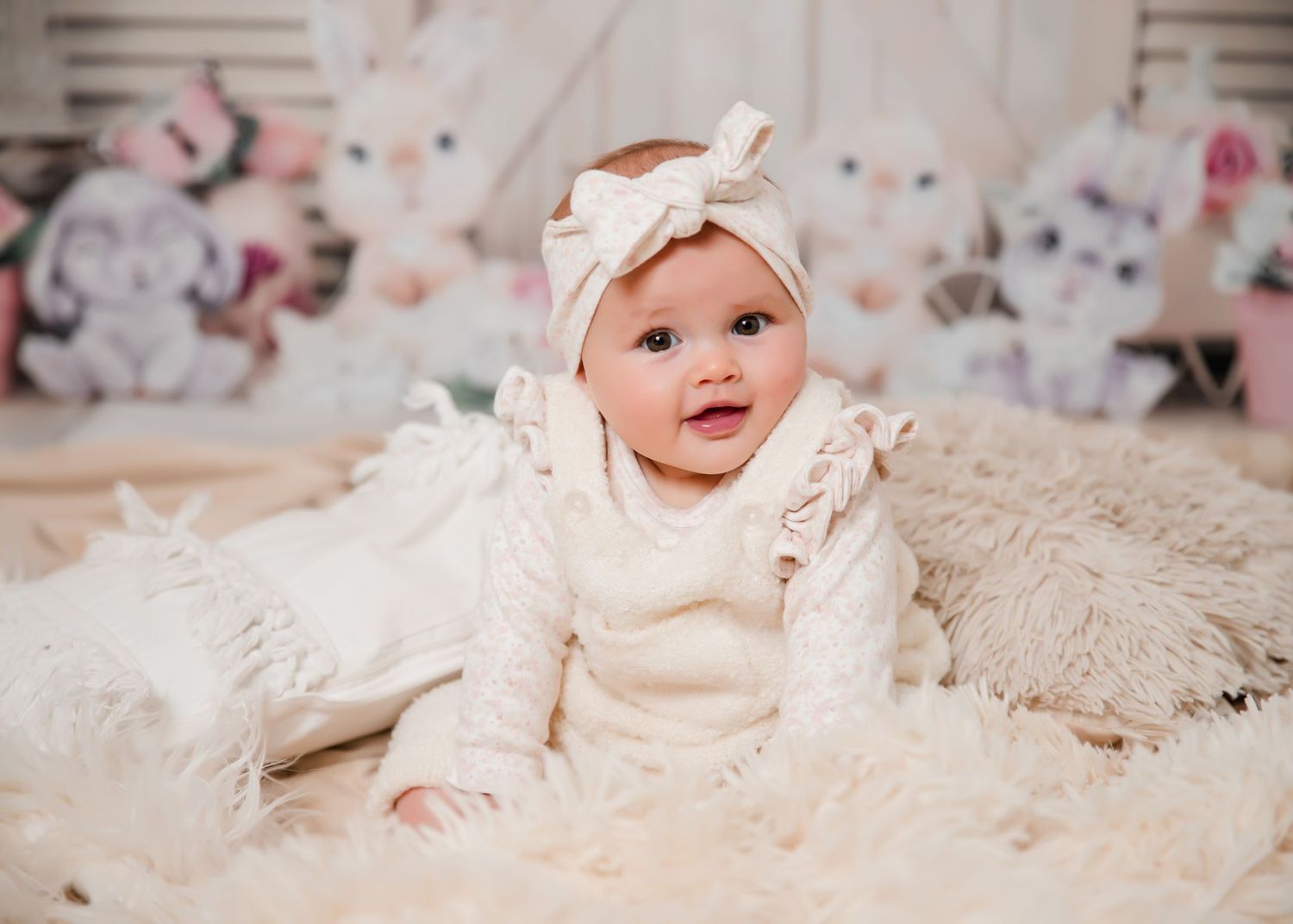 A smiling baby wearing a cream knit sweater and white bow headband sits on a fluffy white rug with plush toys in background.