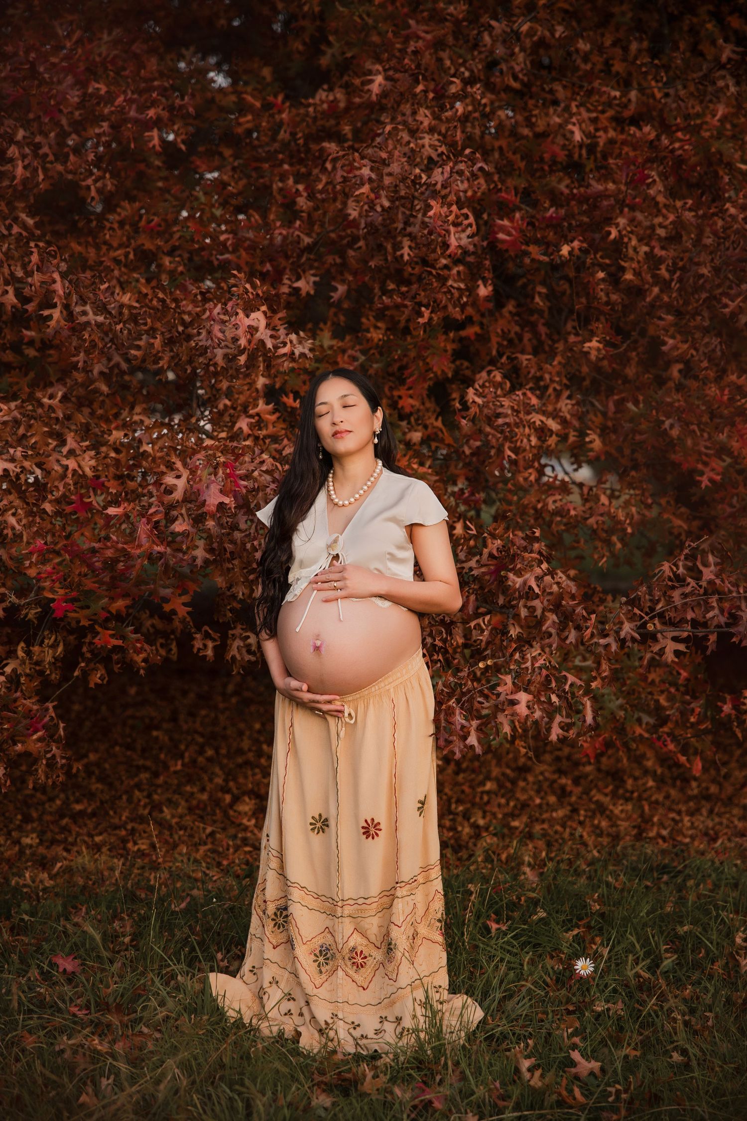 Pregnant woman in beige skirt cradling baby bump outdoors against vibrant red autumn tree leaves.