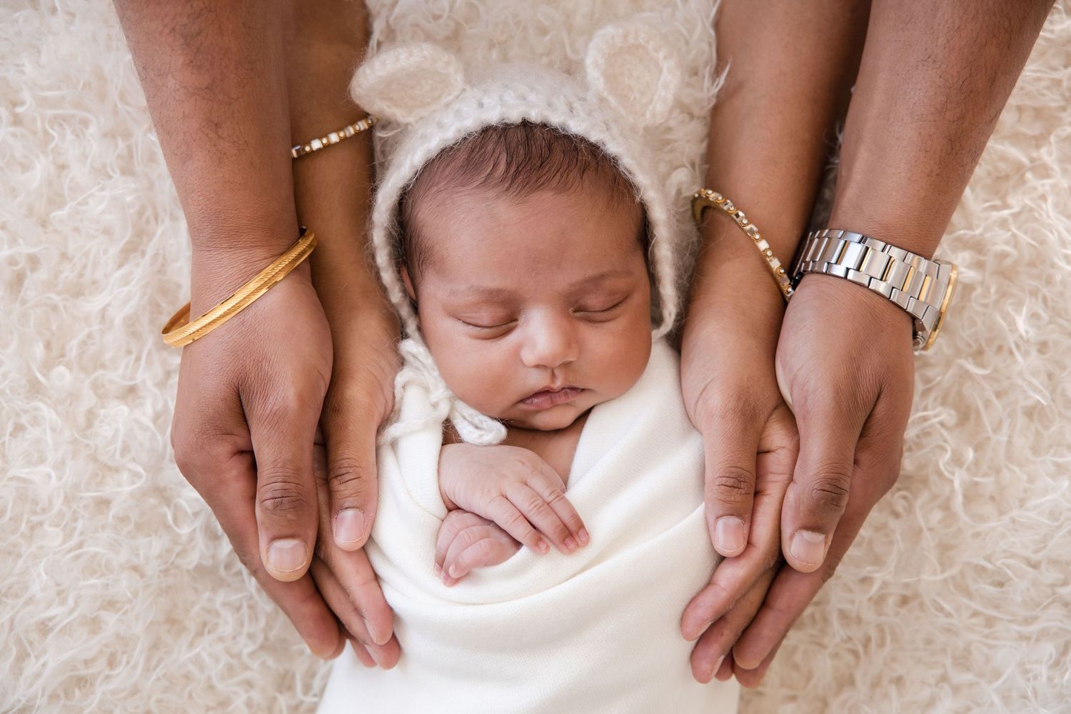 A peaceful sleeping newborn swaddled in white being cradled by gentle hands wearing jewelry on a lace background.