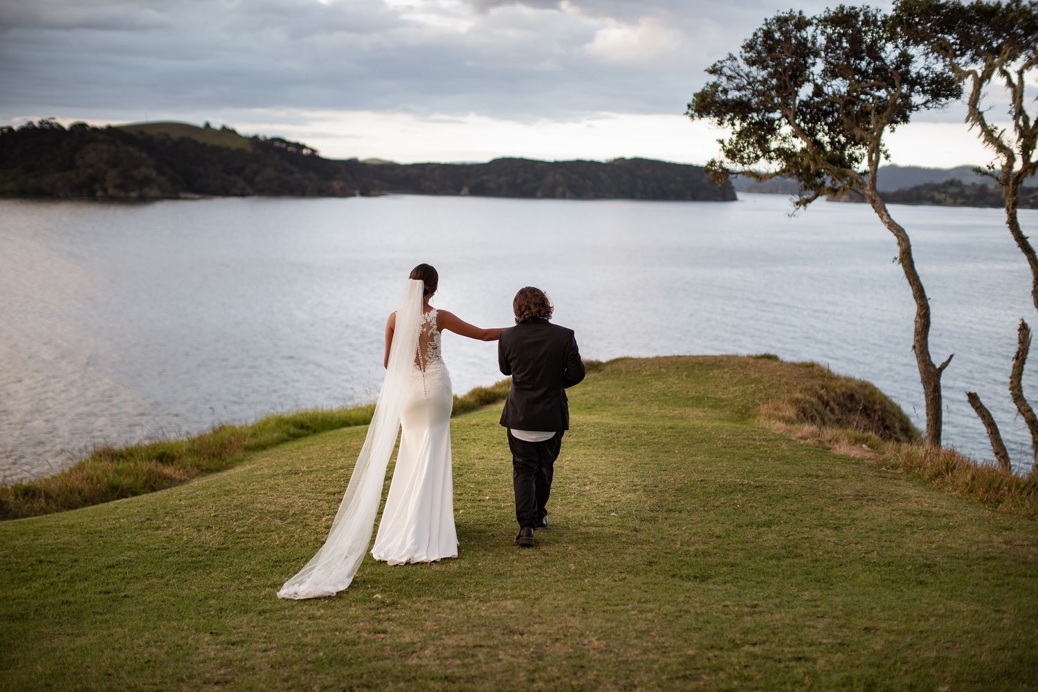 A couple stands together on a grassy cliff overlooking a serene lake on their wedding day.