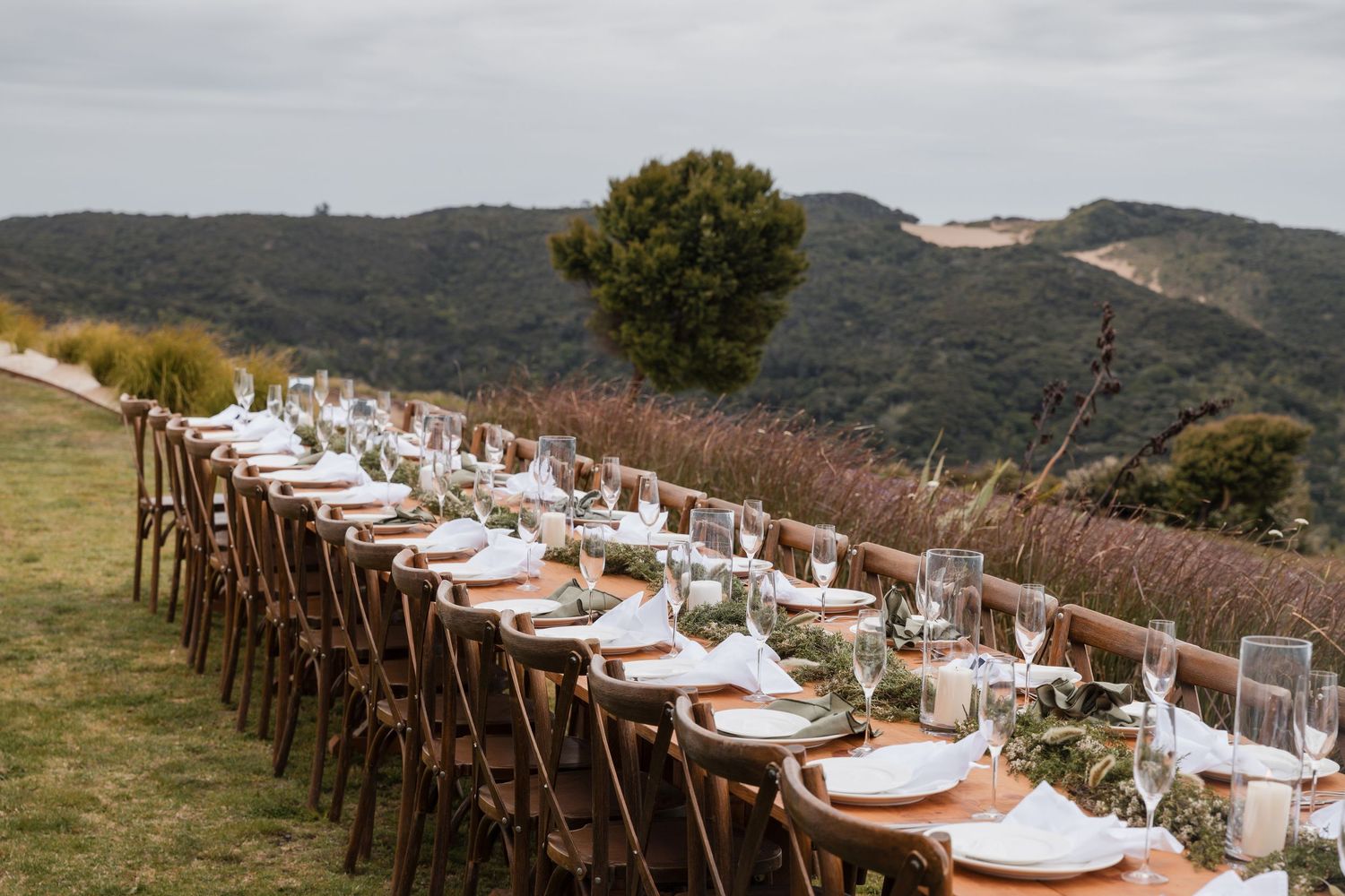 A long rustic outdoor dining table set on a hilltop with wooden chairs and white place settings against mountain views.